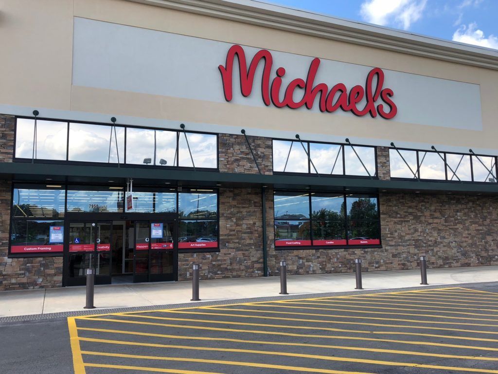 Exterior of a Michaels craft store with a red logo on a stone-facade building and a striped parking lot.
