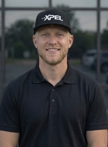 A person smiling, wearing a black collared shirt and a black XPEL baseball cap, standing before a glass building.