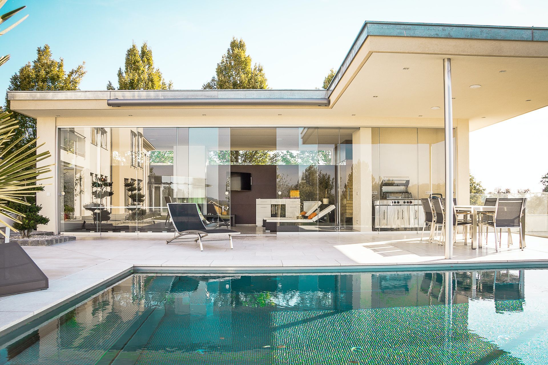 A modern house with floor-to-ceiling glass walls overlooks a shimmering blue swimming pool on a sunny day.