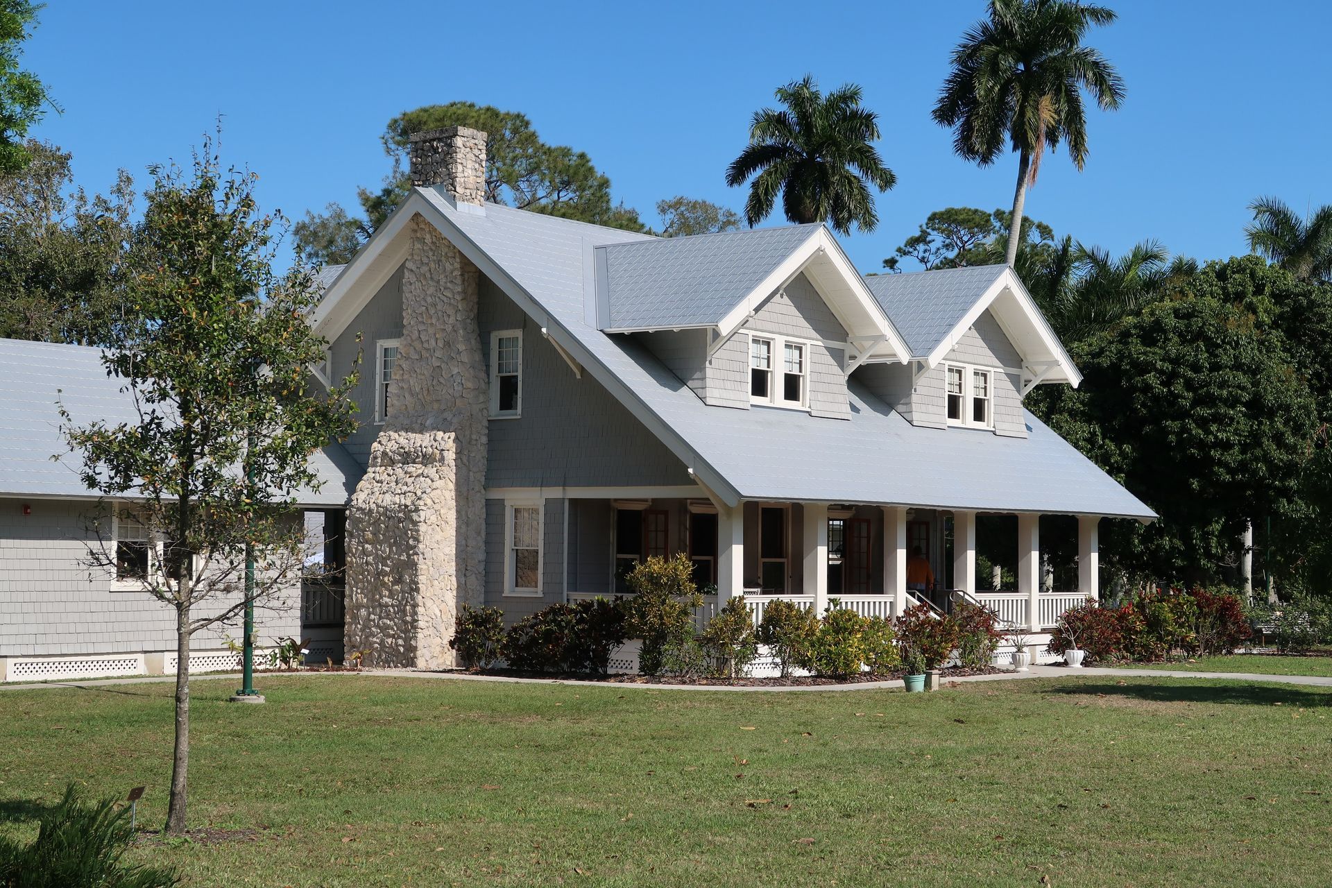 A light gray, two-story house with two front-facing gables, a stone chimney, and a wide porch under a clear blue sky.