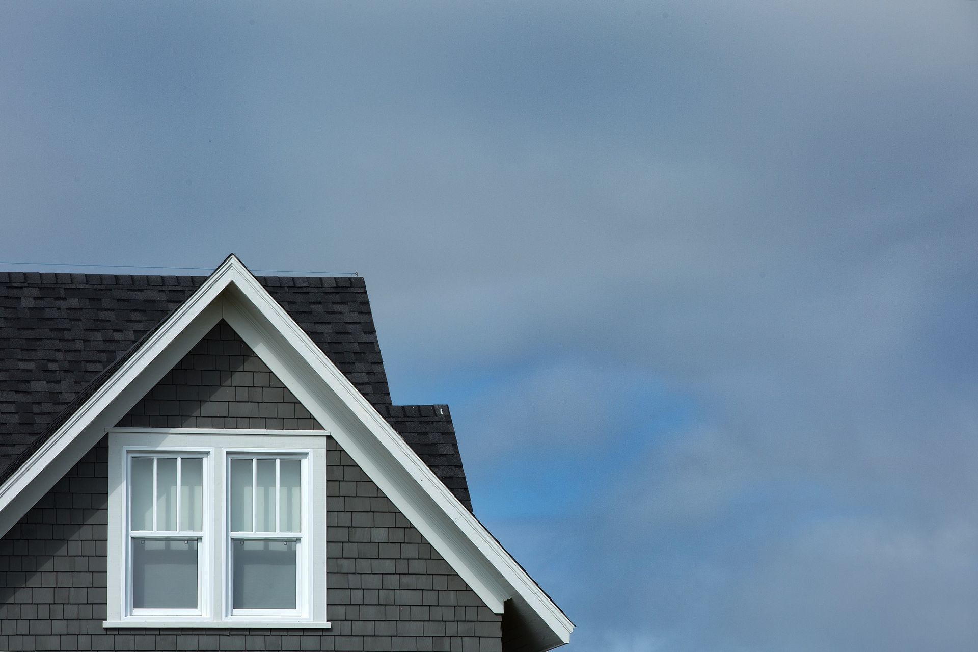 The roof of a gray house with a white window and a cloudy sky in the background.