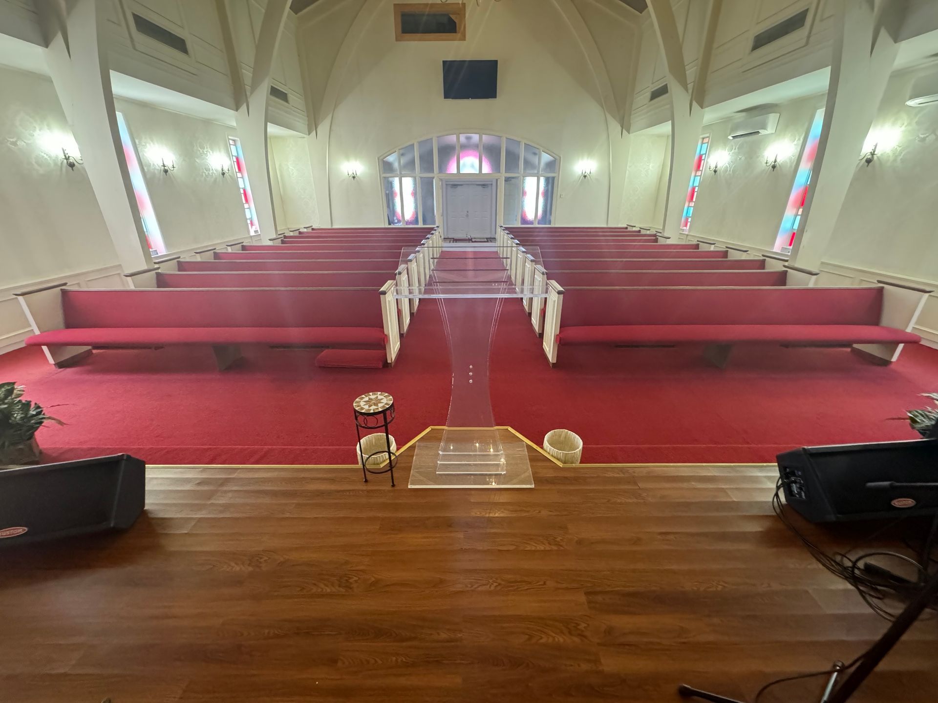 The front of the small chapel. There are 9 rows of classic pews that will seat 100 people comfortably. The carpet is red, there are large stained glass windows letting in bright light. 