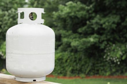 A white propane tank on a wood surface with a green foliage background in an outdoor setting