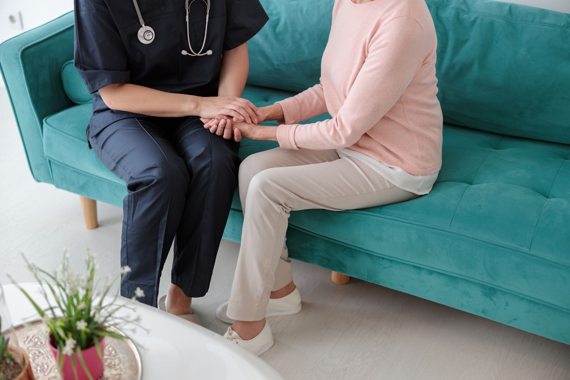 A nurse is holding the hand of an elderly woman while sitting on a couch.
