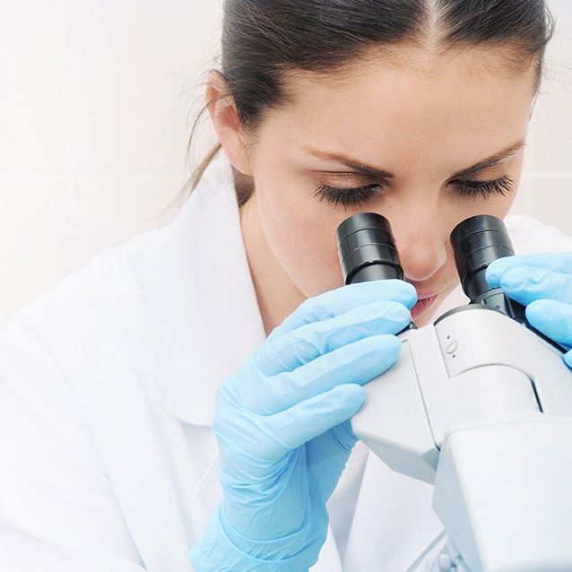 A woman wearing blue gloves is looking through a microscope.