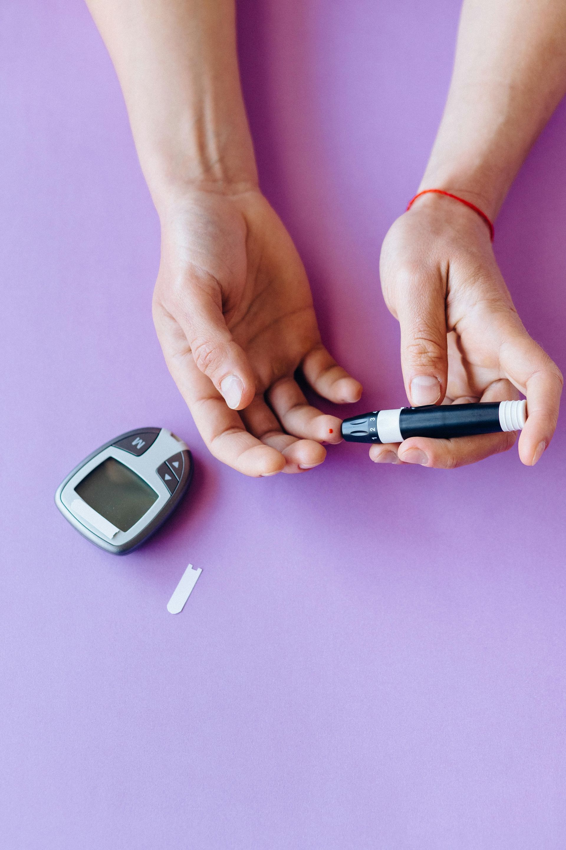 Hands using a lancet to prick a finger for a blood glucose test; meter and test strip on a purple surface.