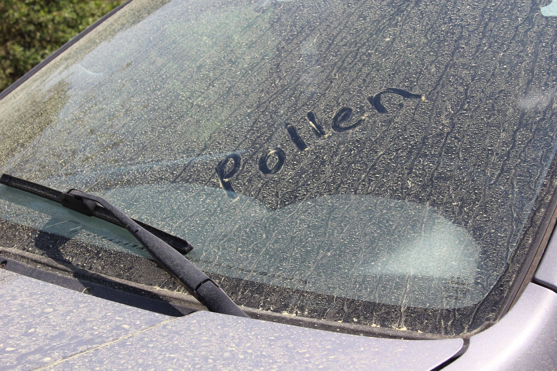 Dirty car windshield with the word “Pollen” written in dust, wiper visible