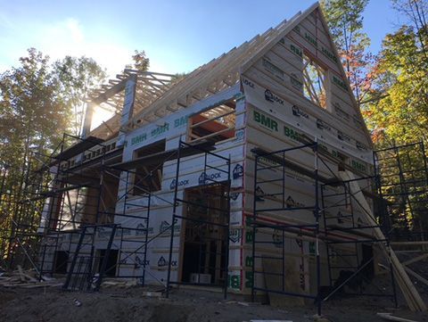 Maison en construction avec échafaudages, ossature bois et bardage. Le soleil brille à travers le toit ouvert.