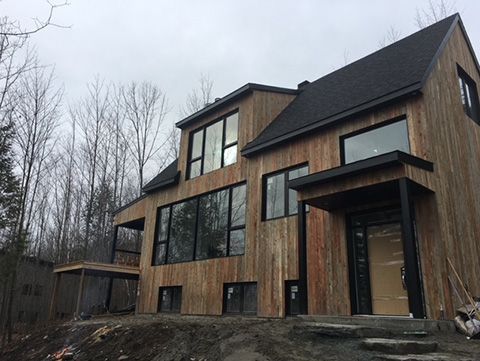 Maison moderne à deux étages avec revêtement en bois brun clair et cadres de fenêtres sombres sous un ciel nuageux.