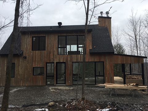 Maison à deux étages en bois avec fenêtres à cadre noir et porche couvert dans une zone boisée.