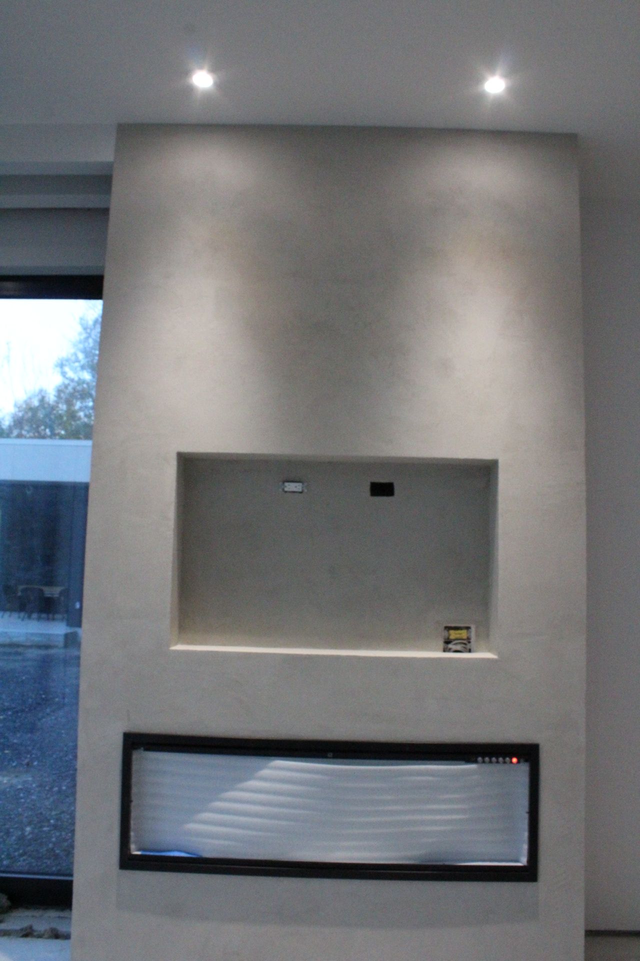 Cheminée encastrée et niche TV sur un mur gris clair, éclairée par des plafonniers.
