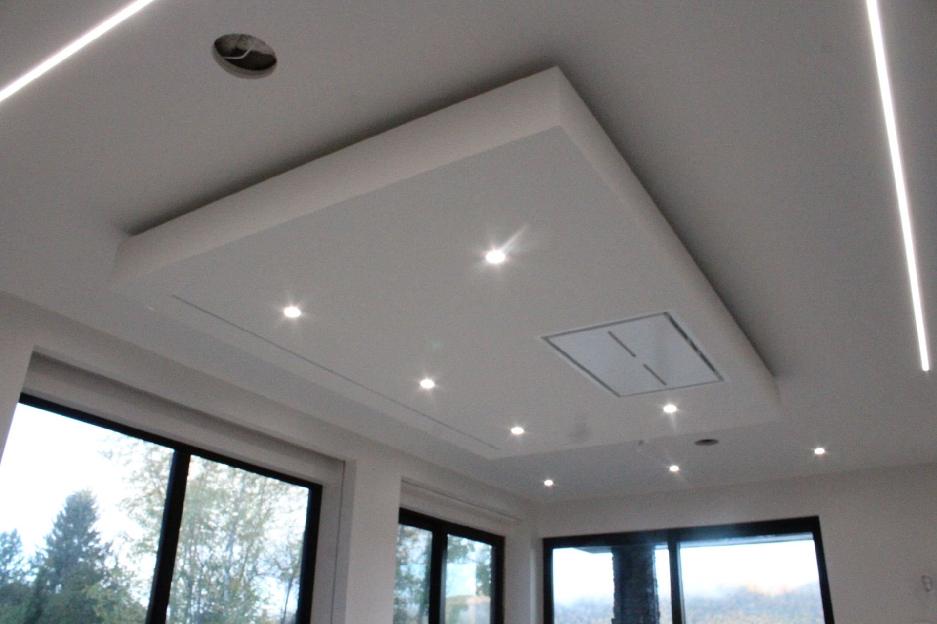 Plafond blanc avec spots encastrés et panneau rectangulaire abritant un évent, fenêtres le long du bas.