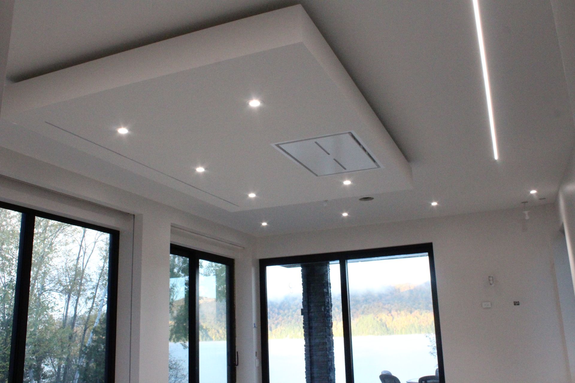 Pièce intérieure lumineuse avec plafond blanc, lumières encastrées et grandes fenêtres donnant sur un paysage.