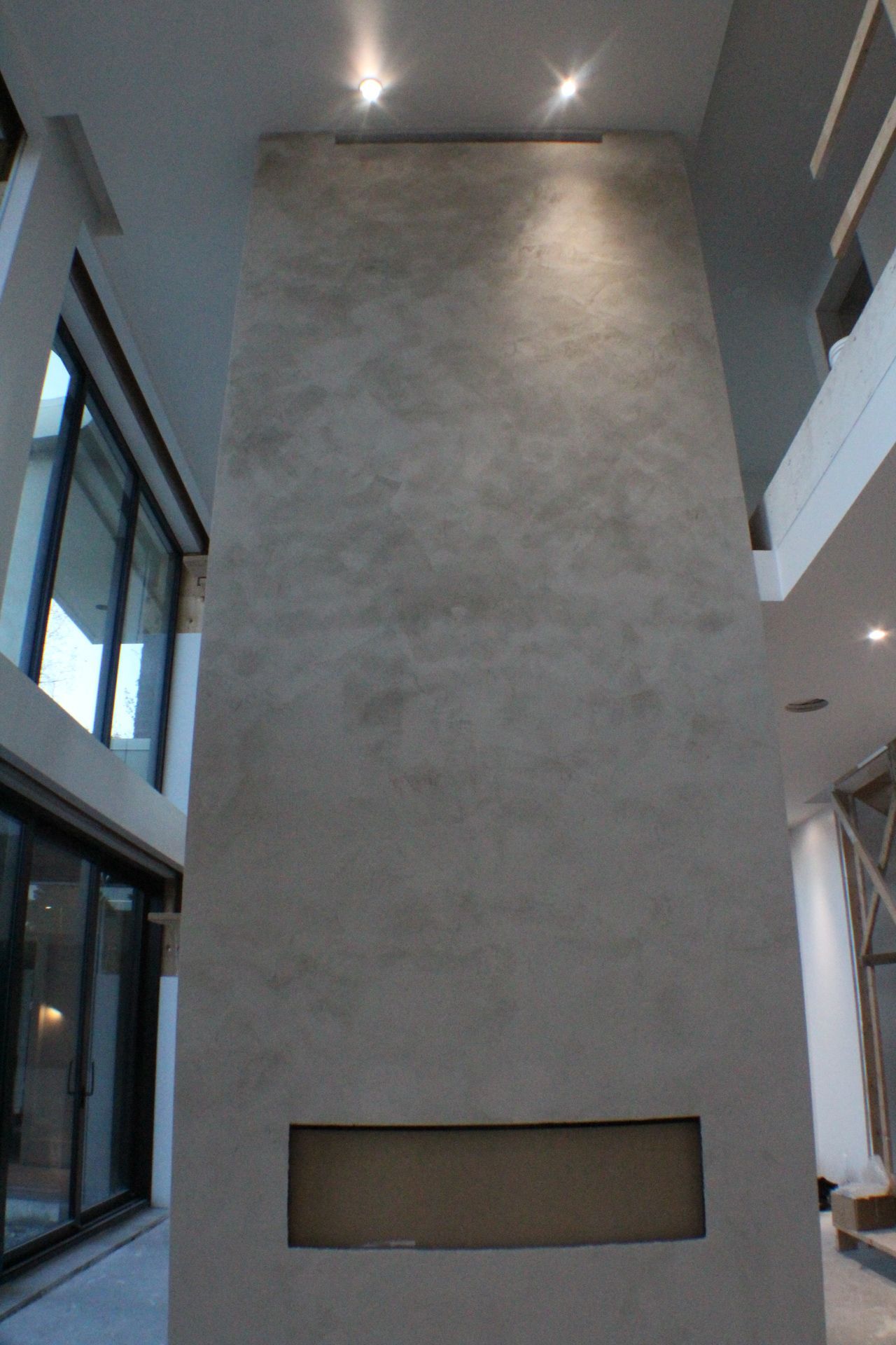 Grand mur texturé gris avec une ouverture rectangulaire encastrée, éclairé par des spots dans une maison moderne.