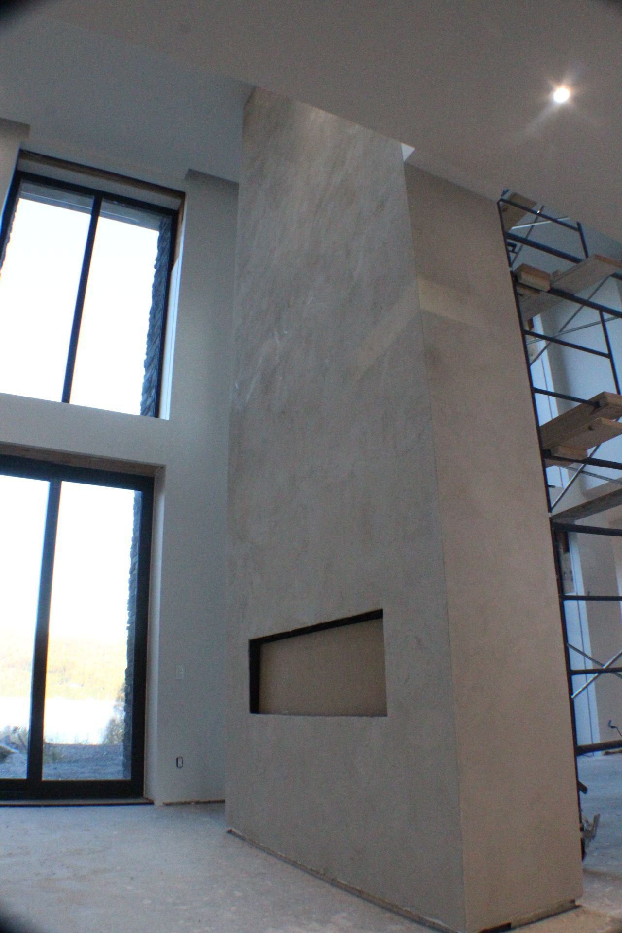 Grande cheminée texturée grise avec insert noir, à côté d'une grande fenêtre et d'un échafaudage dans un espace moderne.