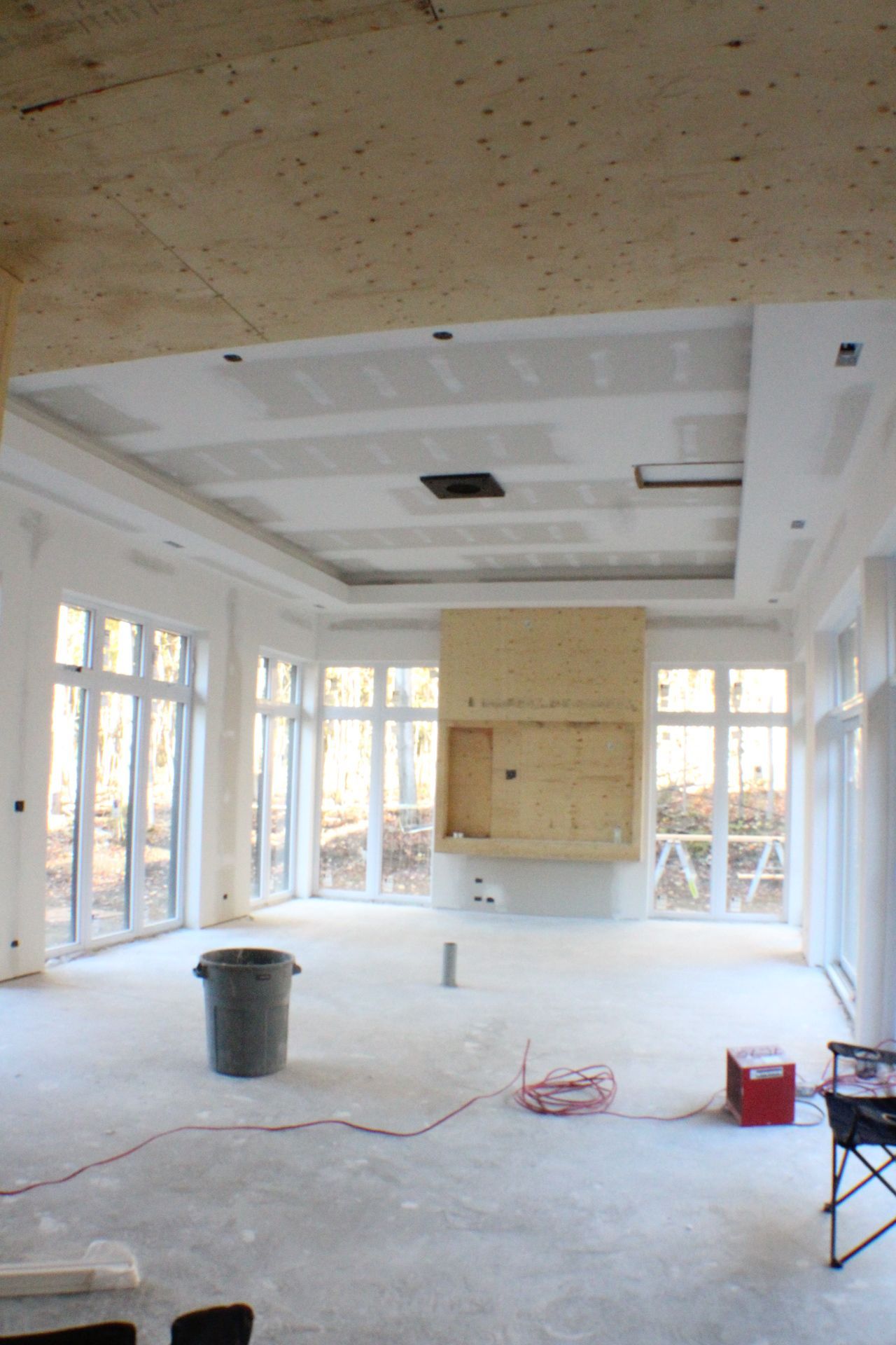 Intérieur en cours de rénovation : plafond en plaques de plâtre avec éclairage encastré, fenêtres et cheminée. Sol gris et poubelle.