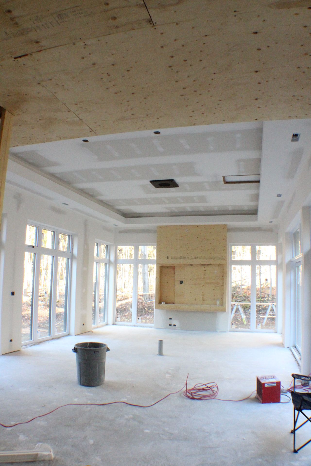 Intérieur d'une maison en construction avec cheminée, fenêtres et surfaces non finies. Une poubelle et des outils sont posés au sol.