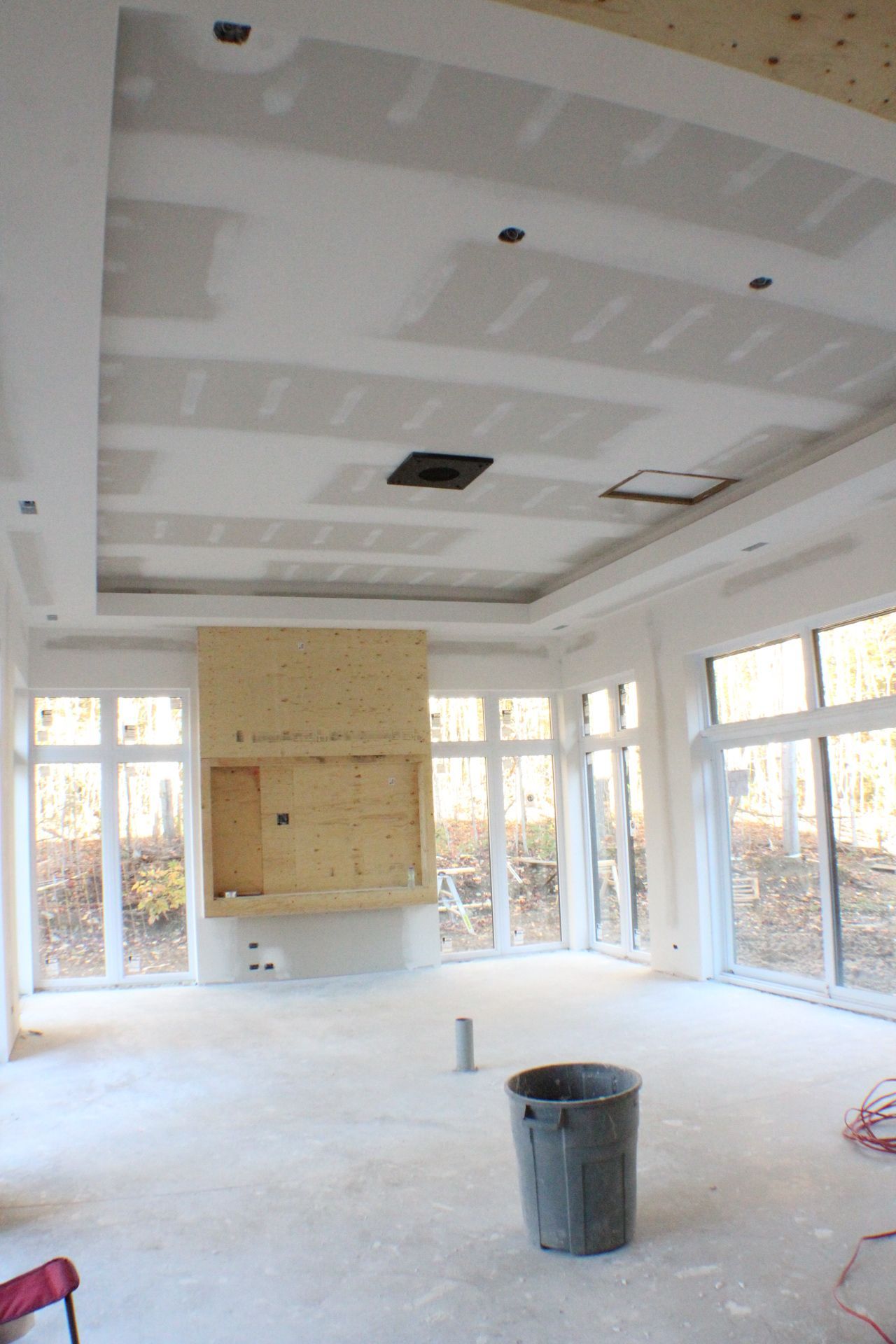 Intérieur en construction avec cheminée et grandes fenêtres; poubelle grise posée sur le sol en béton.