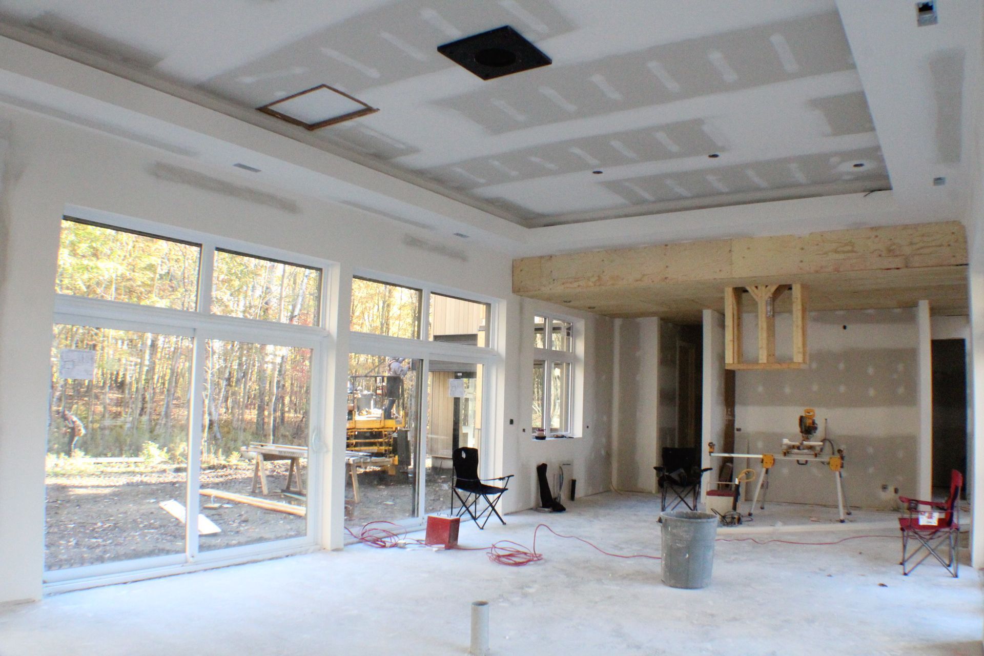 Intérieur d'une pièce en construction avec de grandes fenêtres, des cloisons sèches et des poutres apparentes, contenant des outils et des matériaux.