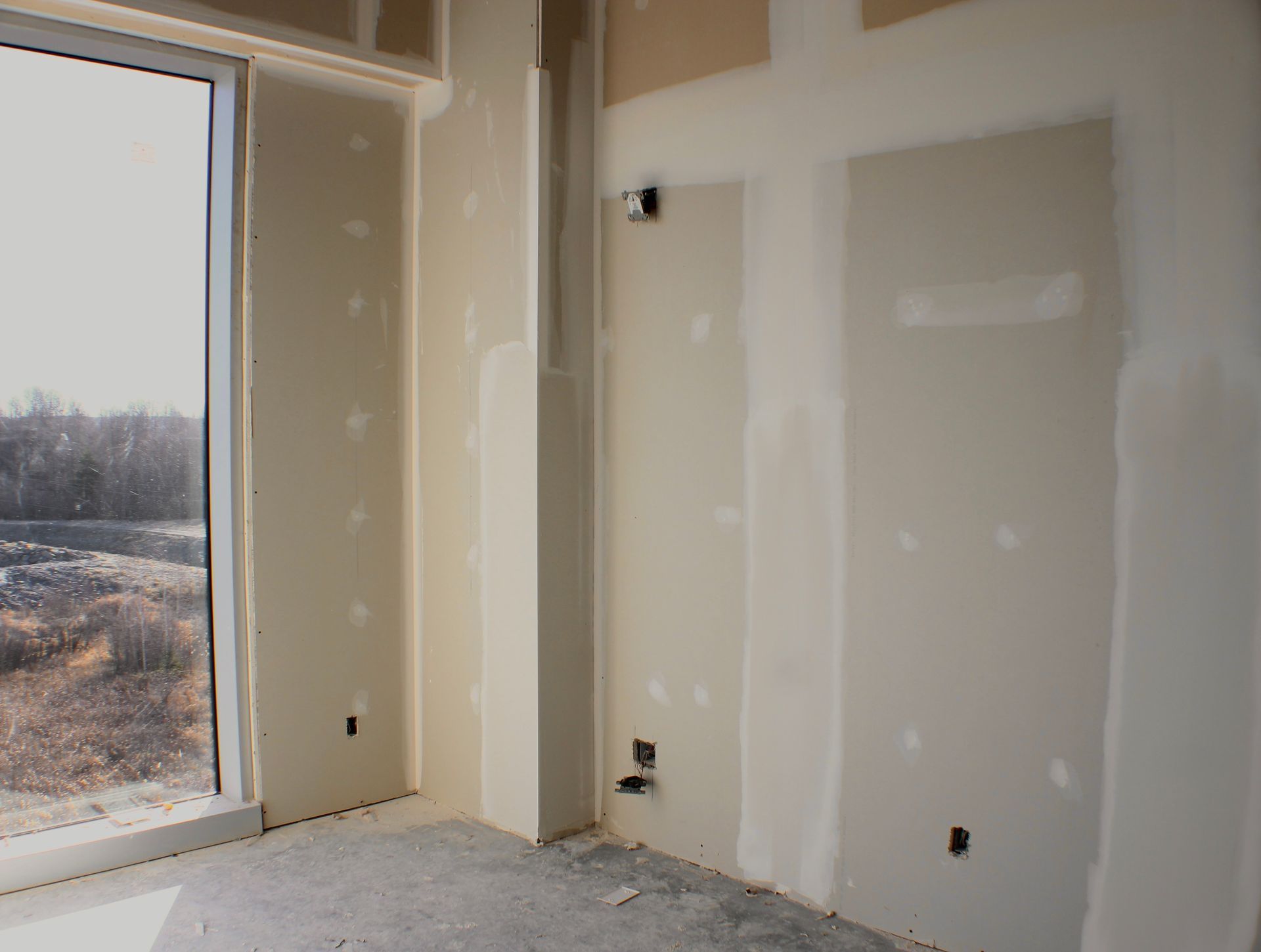 Intérieur d'une pièce en construction, avec des cloisons sèches sur les murs, une fenêtre et des boîtiers électriques exposés.