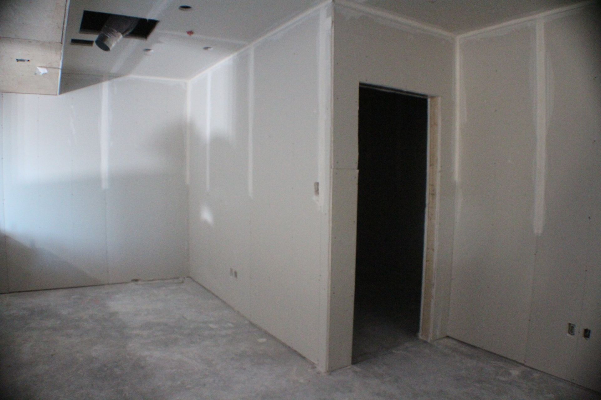 Vue intérieure d'une pièce en construction avec murs en plaques de plâtre, porte ouverte et sol en béton brut.