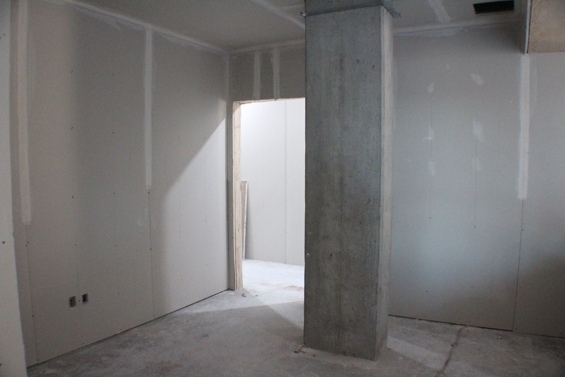 Intérieur avec murs recouverts de plaques de plâtre, colonne en béton et porte. Construction inachevée avec sol en béton brut.
