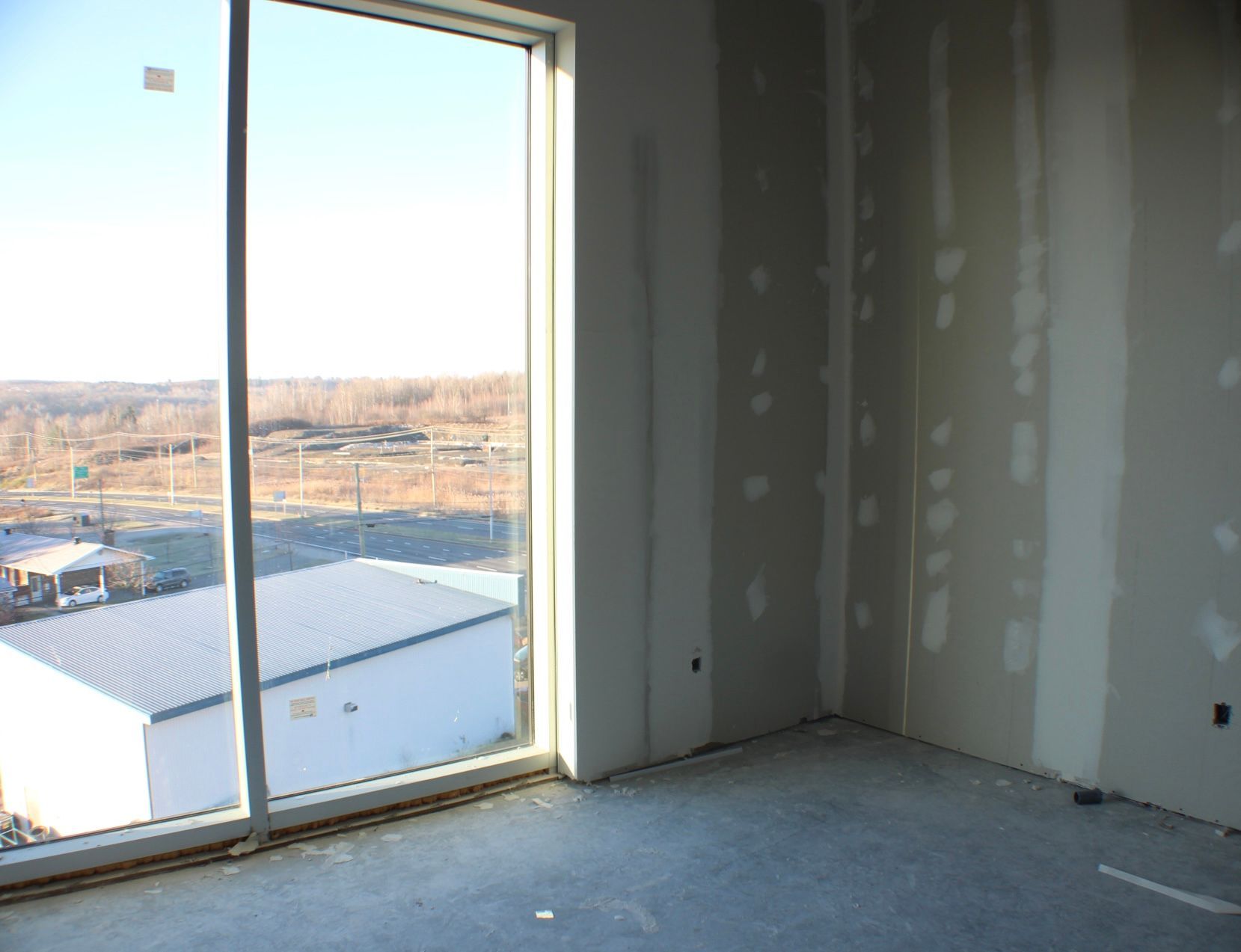 Vue intérieure d'une pièce en construction, avec une grande fenêtre et des cloisons sèches non finies.