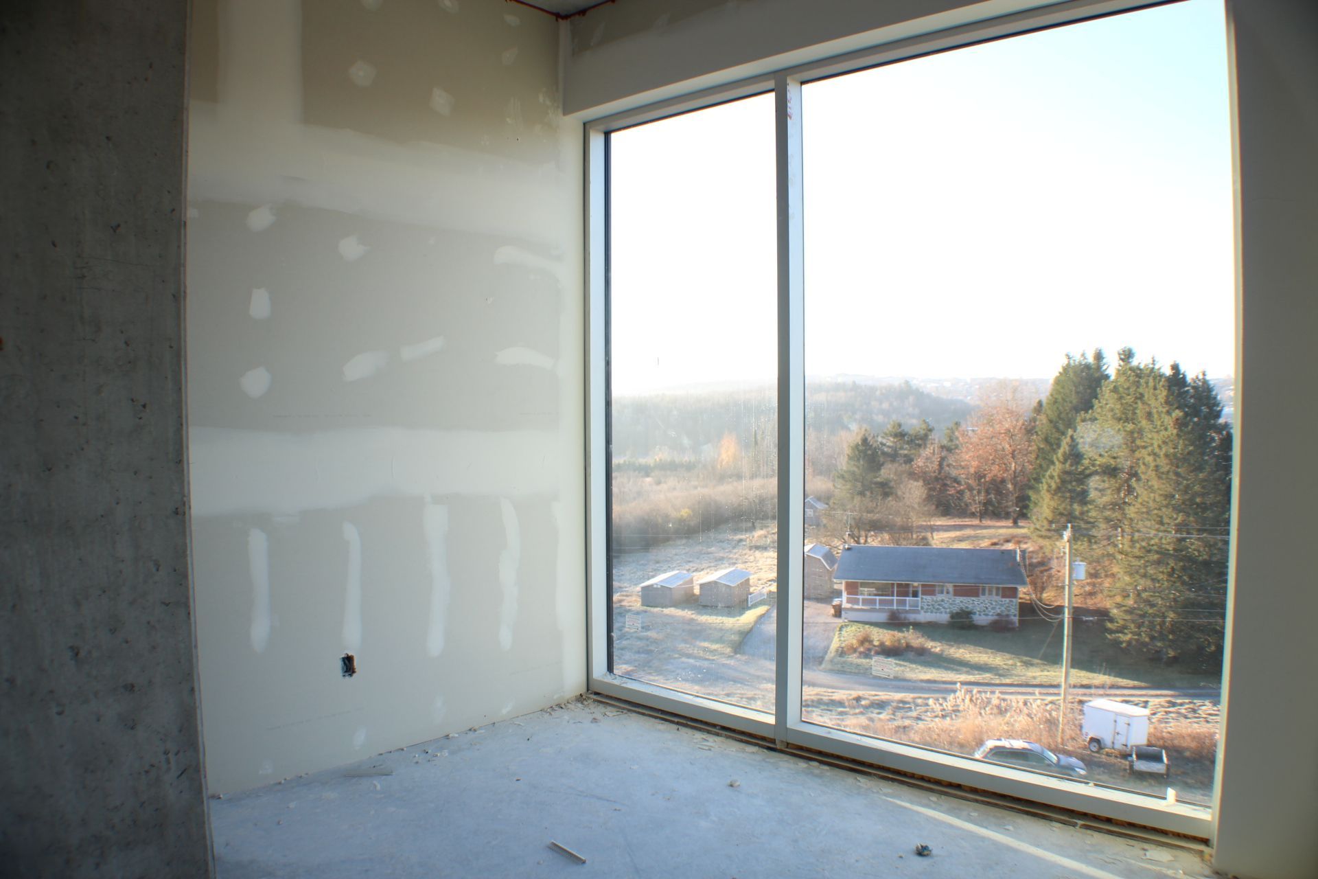 Vue intérieure d'une pièce avec des cloisons sèches non finies et une grande fenêtre donnant sur un paysage rural.