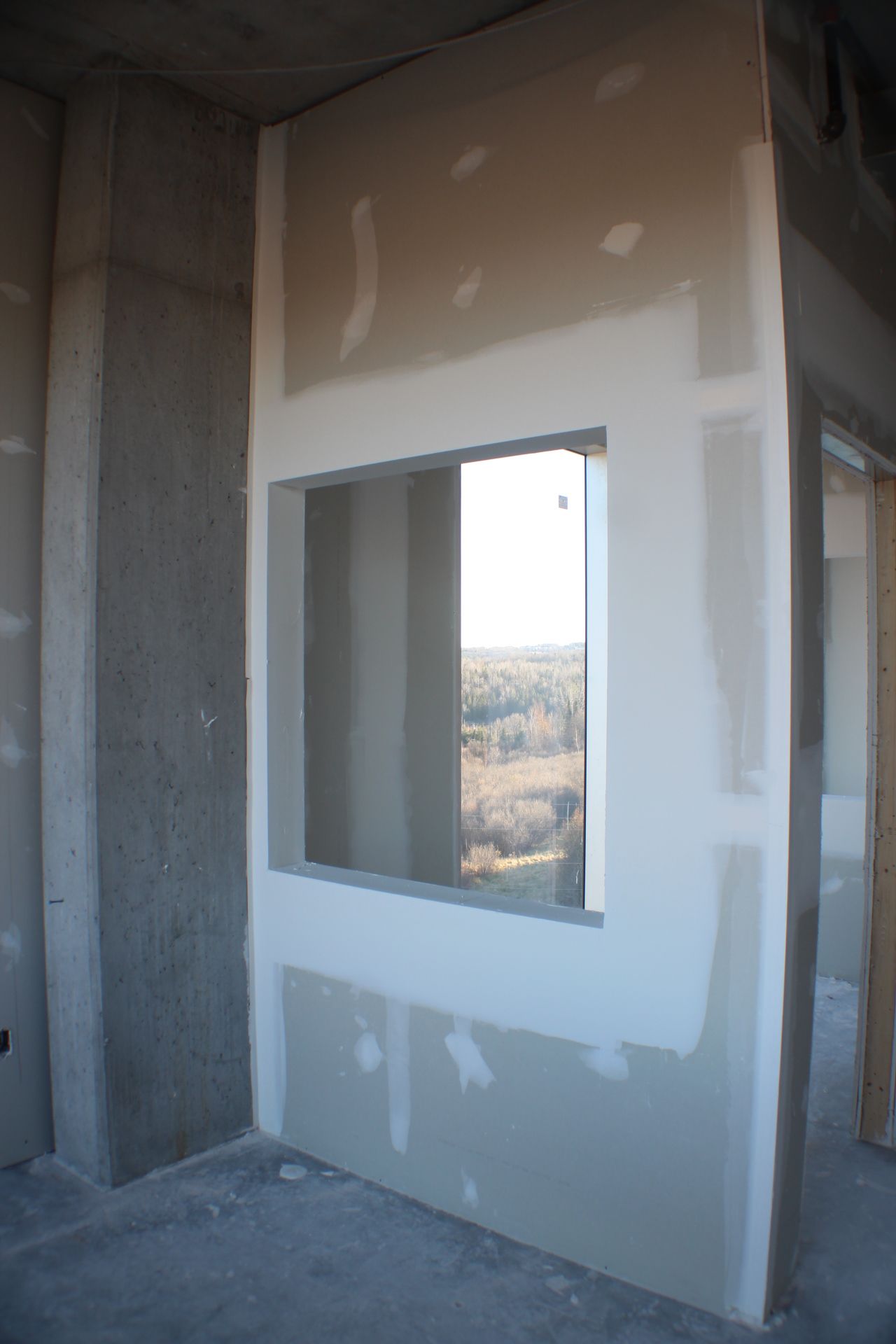Vue intérieure d'une pièce en construction, montrant une fenêtre encadrée de cloisons sèches donnant sur une ligne d'arbres lointaine.