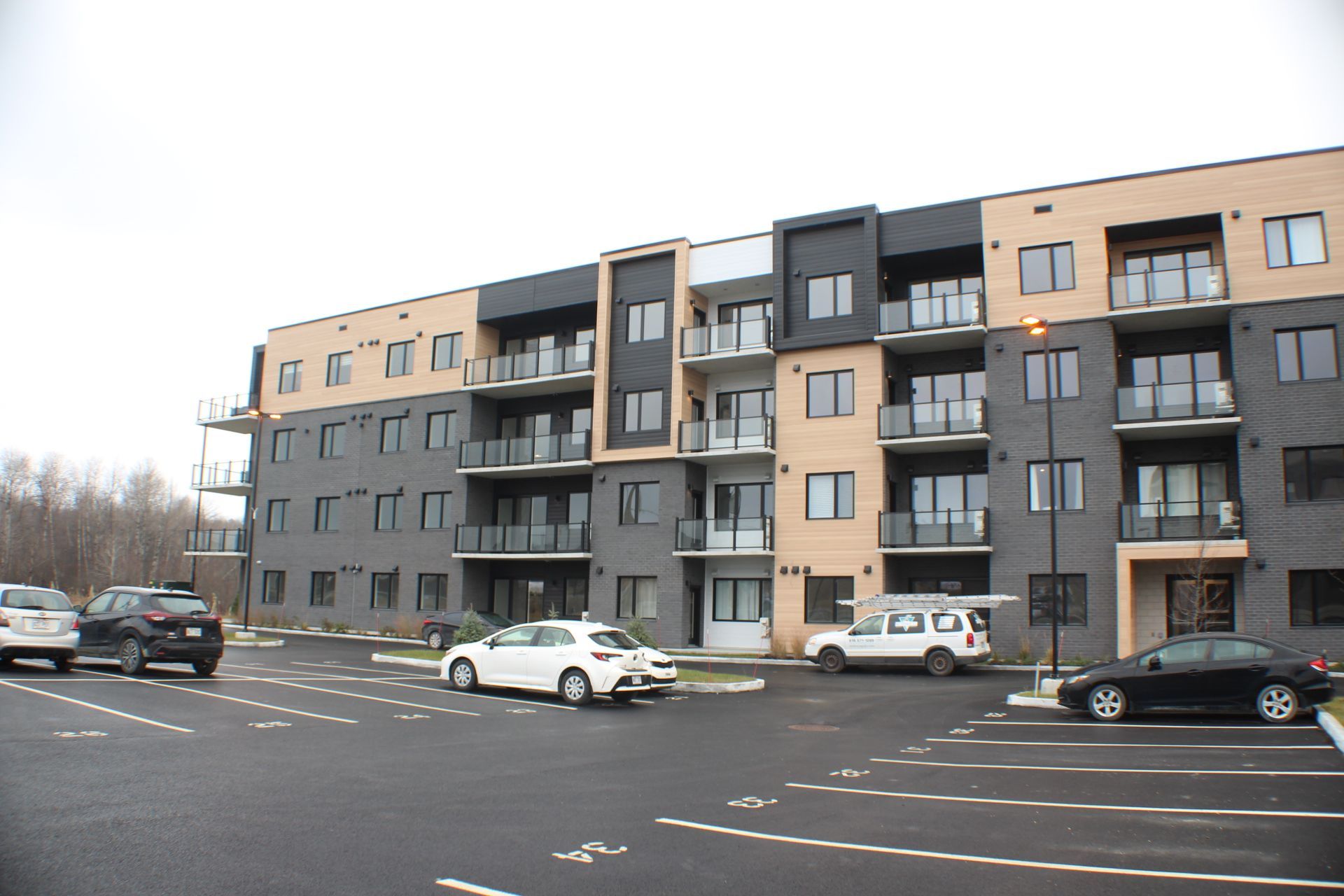 Immeuble d'appartements moderne à plusieurs étages avec balcons, voitures garées devant et ciel couvert.