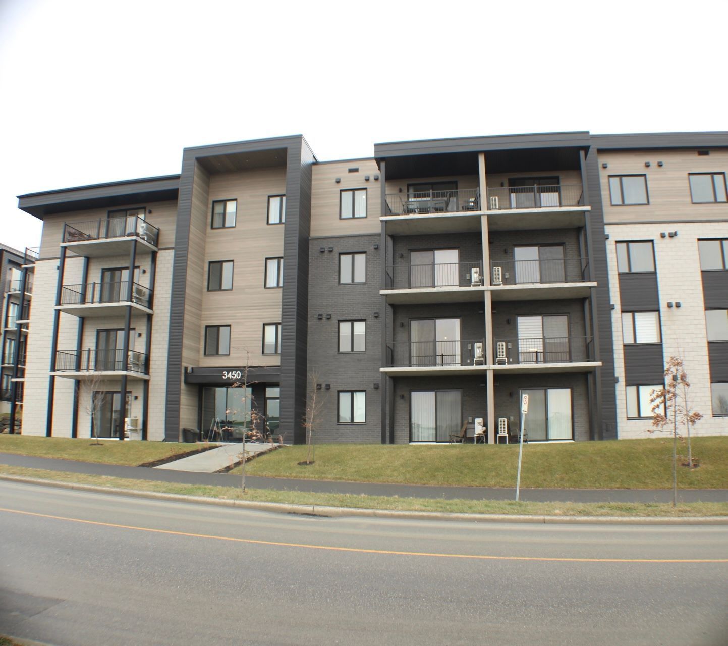 Immeuble d'habitation avec balcons, panneaux clairs et foncés, en retrait d'une rue sur une légère colline.
