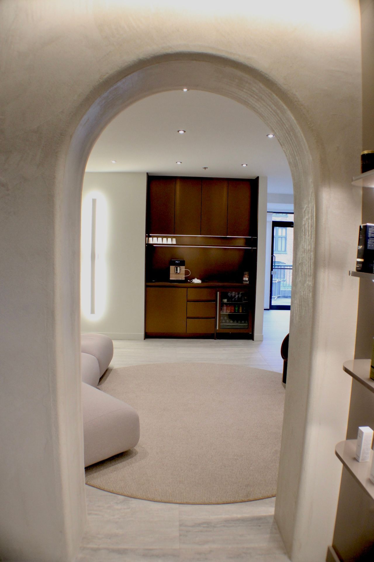 Vue sous arcade d'une salle d'attente aux tons neutres : canapé, tapis, armoires, mini-réfrigérateur.