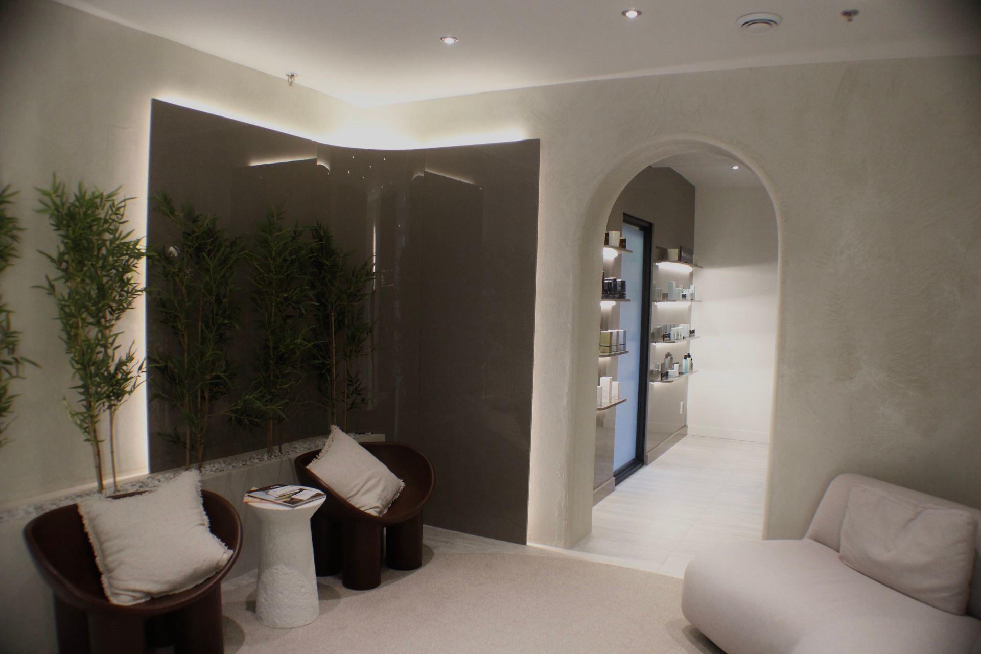 Une salle d'attente dans un spa : deux chaises, des plantes, des couleurs neutres, une porte en arc et une chaise longue.