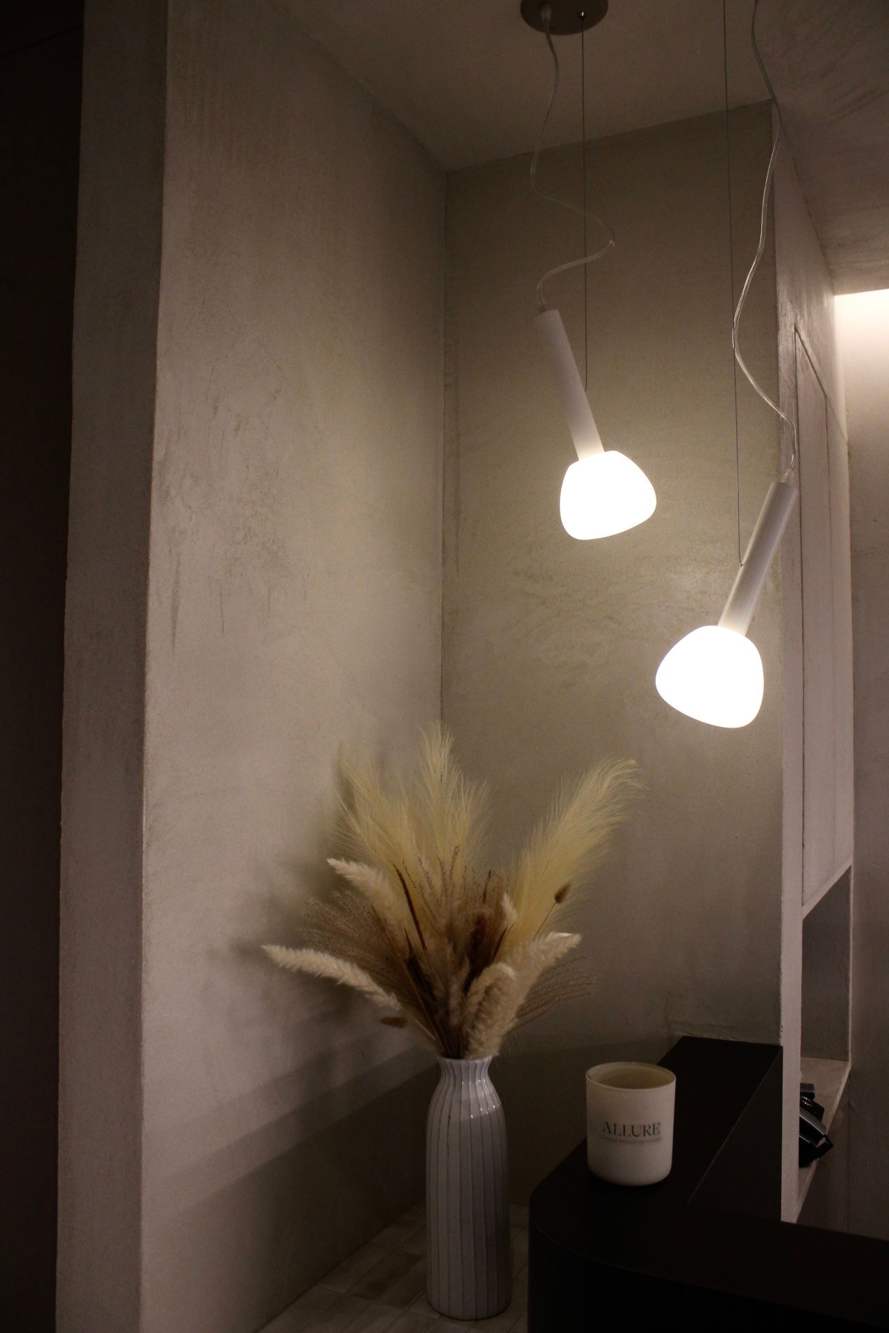 Coin avec de l'herbe de la pampa dans un vase, éclairé par deux suspensions, avec une bougie sur une surface sombre.