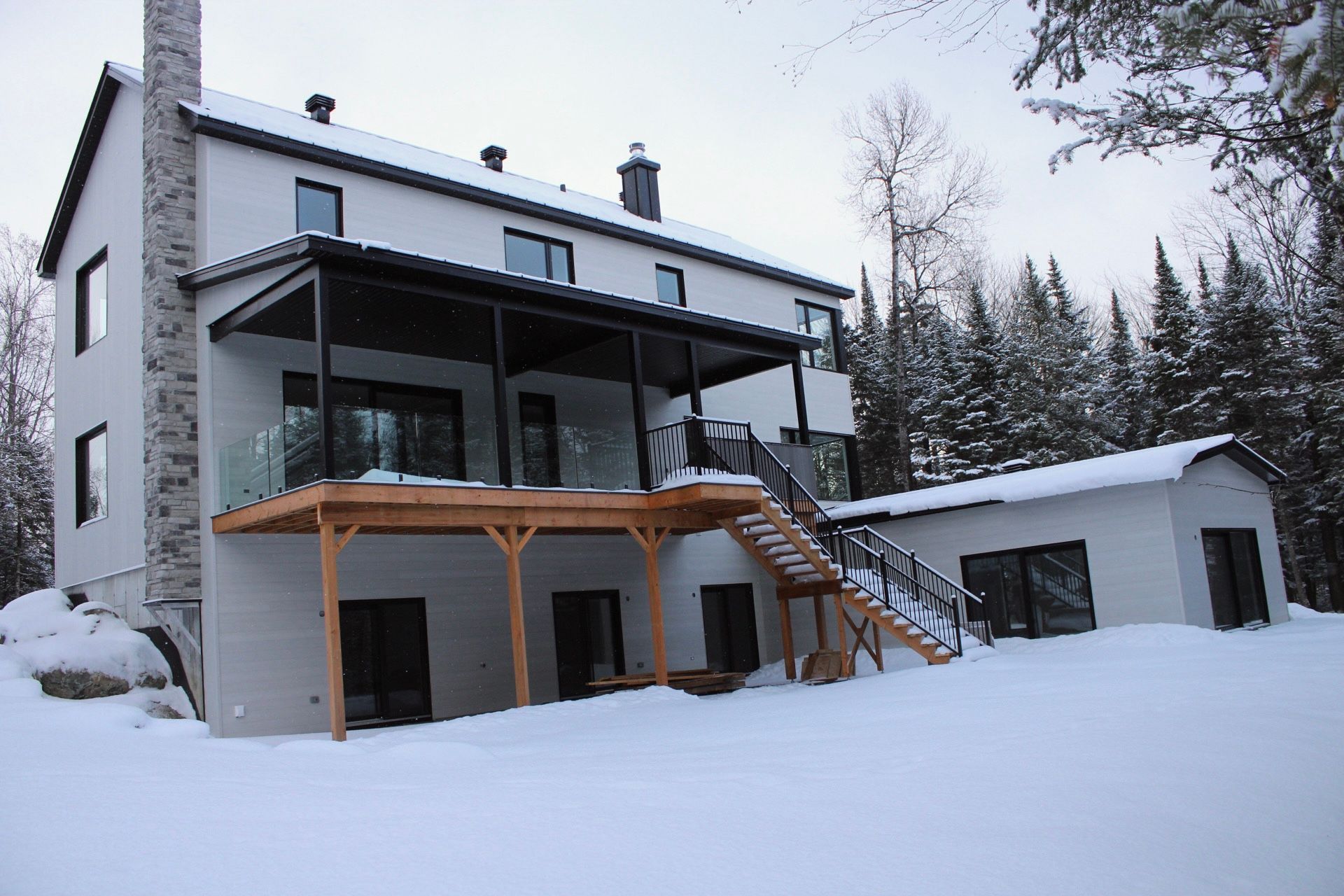 Maison à deux étages avec terrasse et escalier recouverts de neige, entourée d'arbres; ciel couvert.