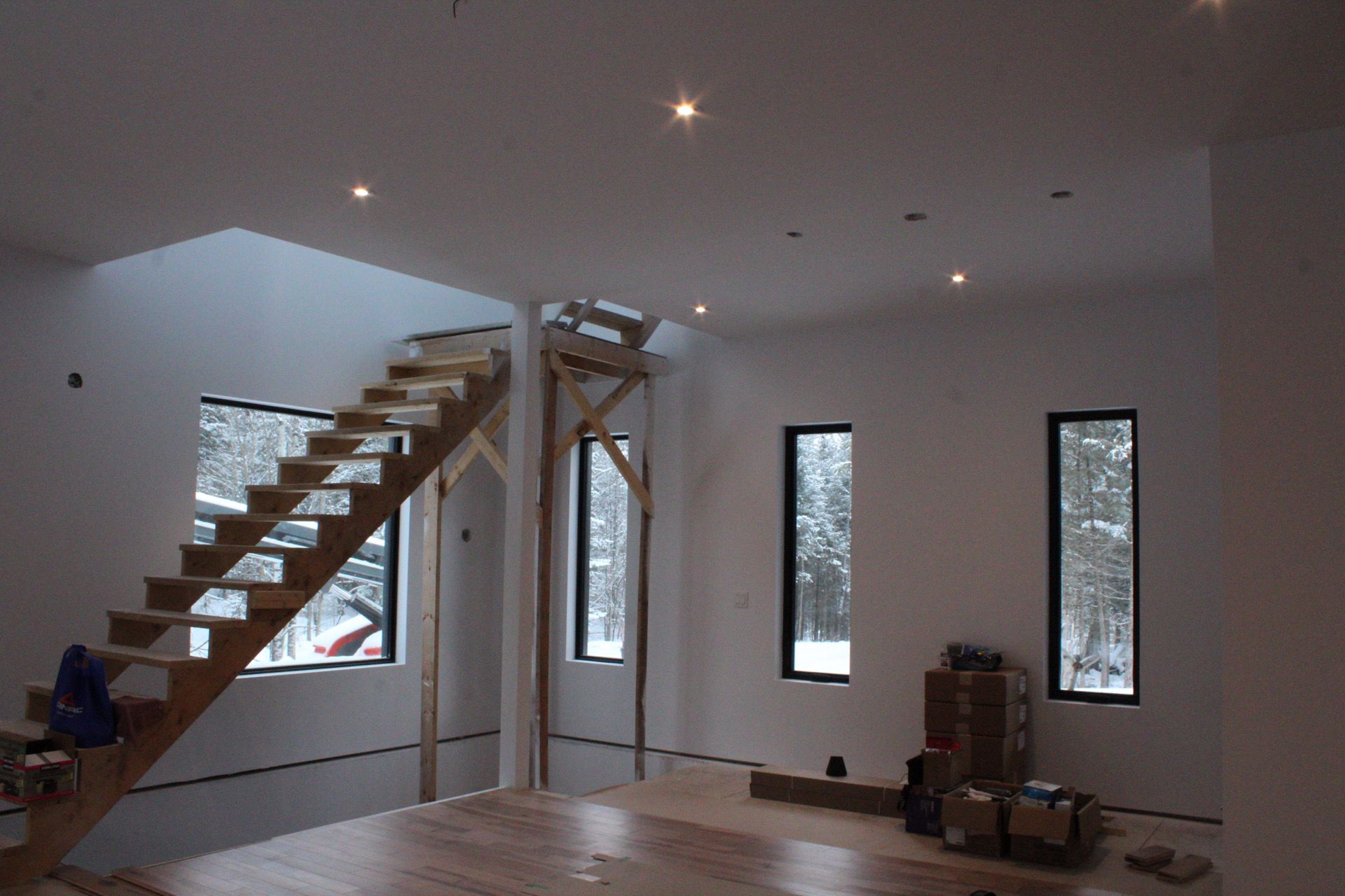 Intérieur d'une maison en construction : escalier en bois, fenêtres étroites, éclairage encastré, caissons au sol, murs blancs.