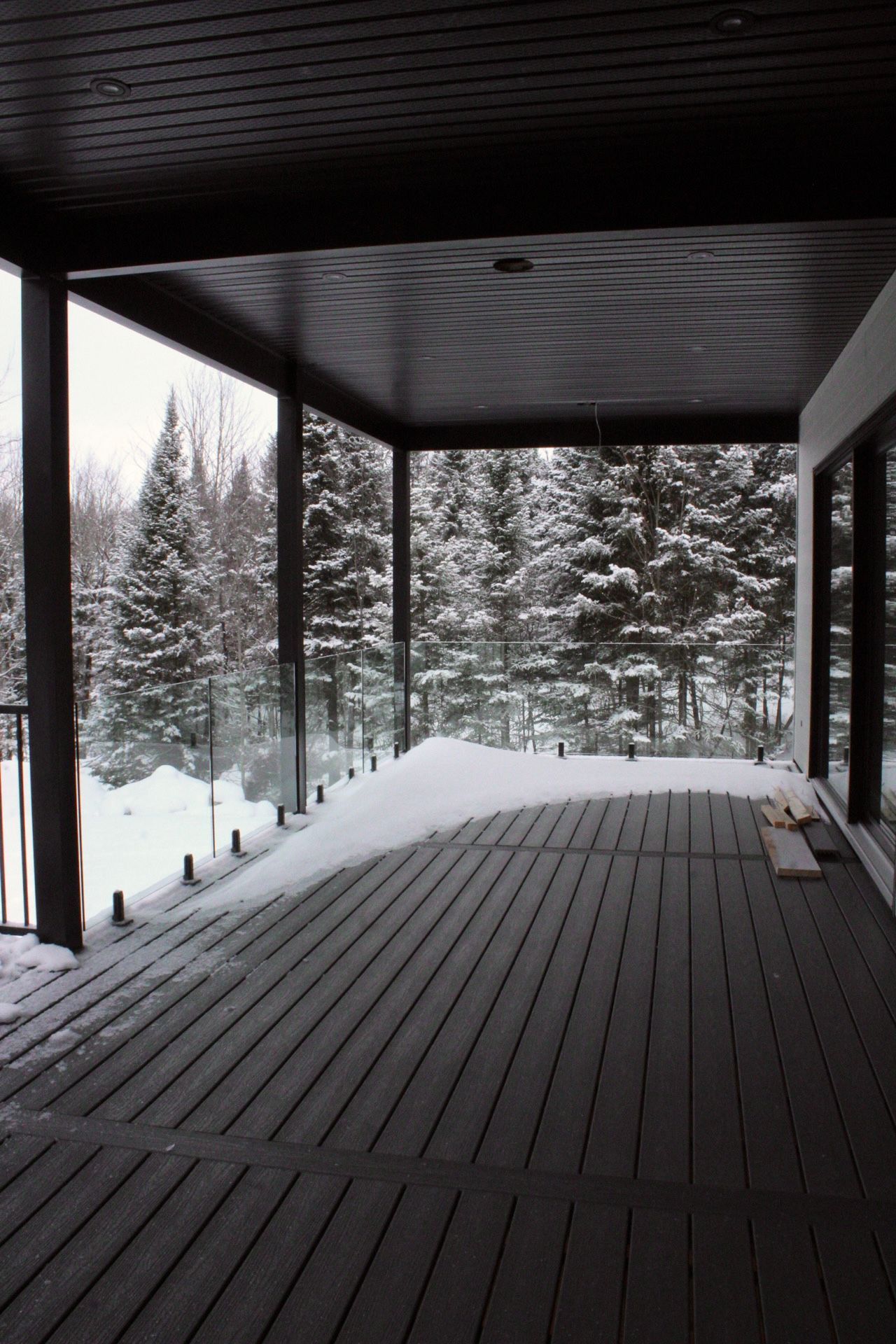 Terrasse couverte avec paysage enneigé en hiver. Planches sombres, garde-corps en verre, arbres enneigés en arrière-plan.