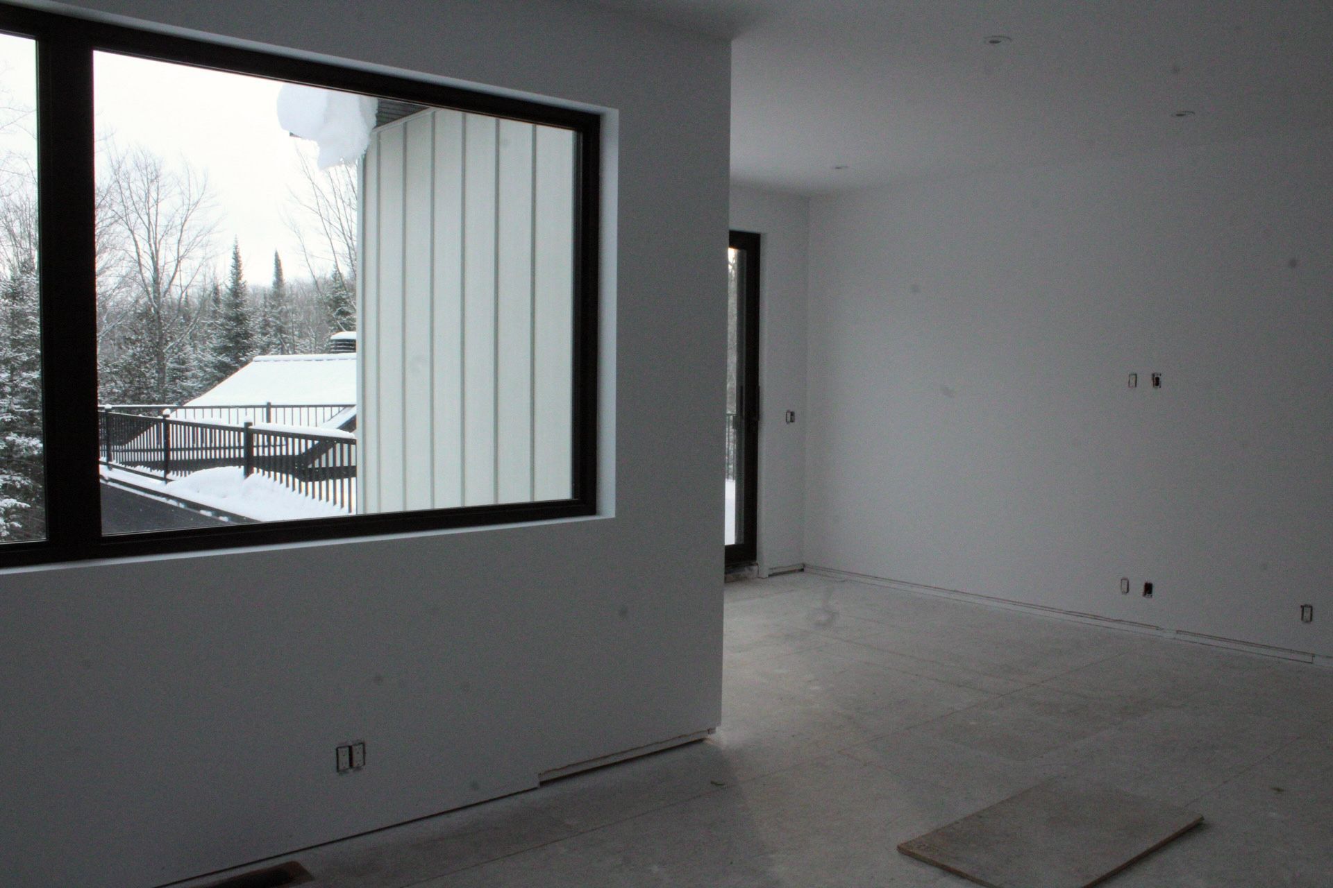 Une pièce vide aux murs blancs avec une grande fenêtre donnant sur une scène extérieure enneigée ; cadre de fenêtre noir.