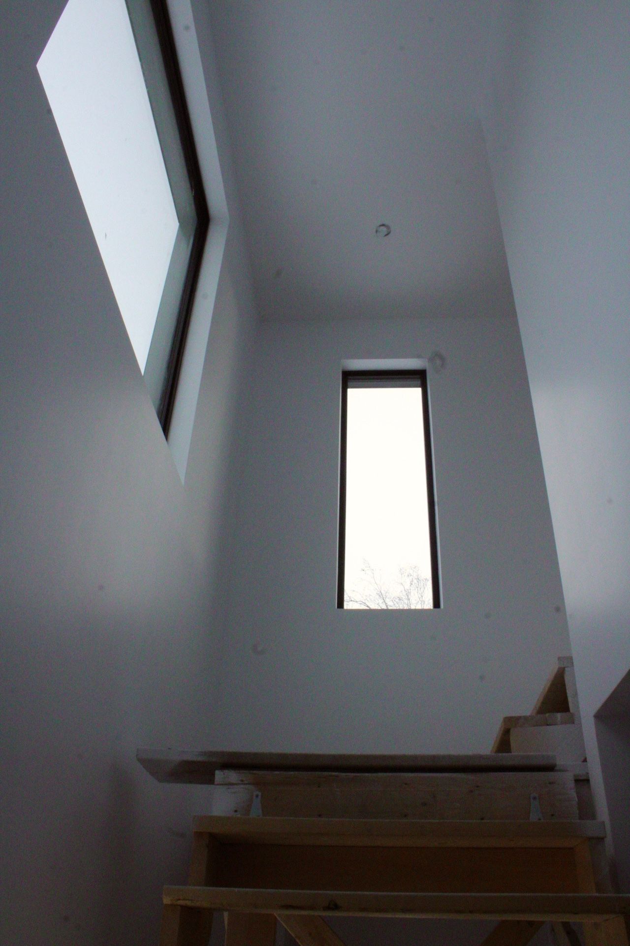 Escalier en bois menant vers le haut avec deux fenêtres, laissant entrer la lumière naturelle, dans un espace aux murs blancs.