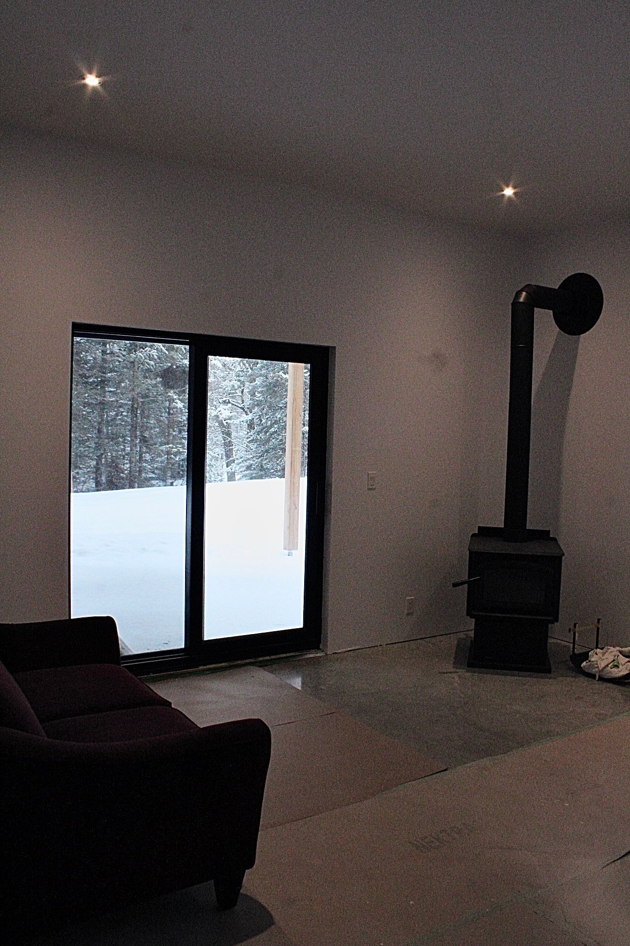 Un intérieur sombre avec une cheminée, une porte coulissante en verre donnant sur l'extérieur enneigé et un canapé.