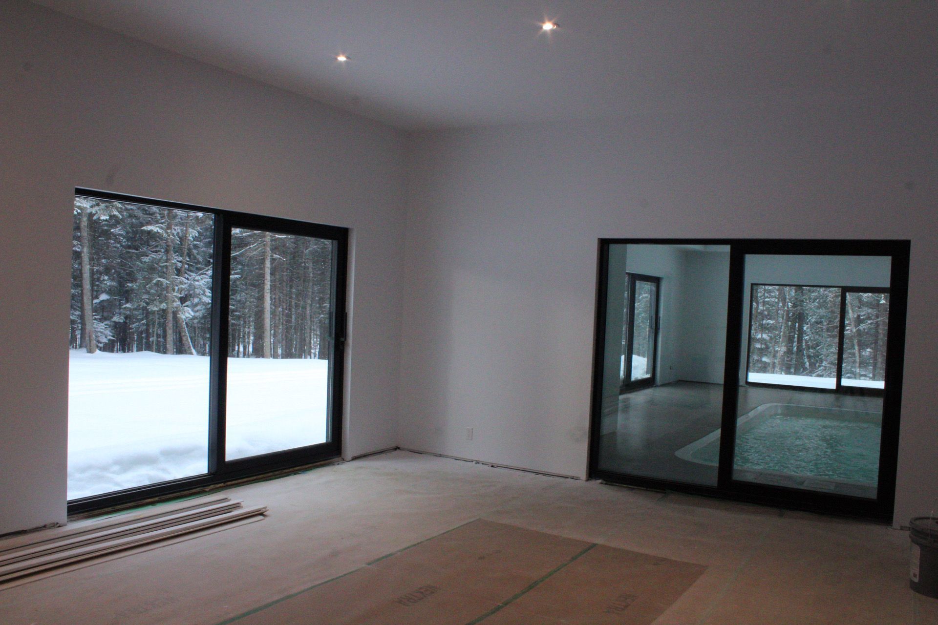Pièce vide avec deux grandes fenêtres à cadre noir donnant sur une forêt enneigée.