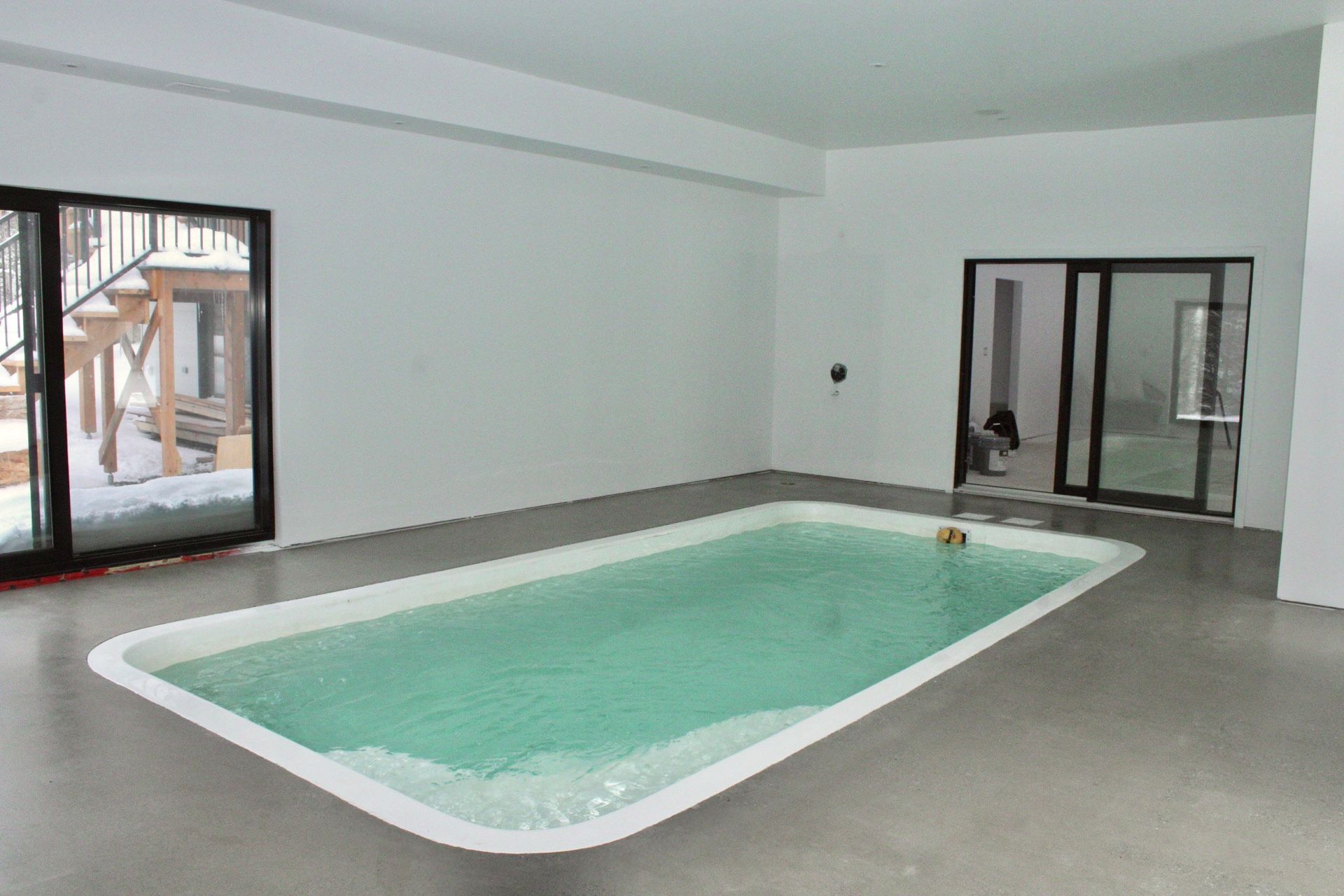 Piscine intérieure rectangulaire avec eau turquoise, murs blancs et grandes fenêtres à cadre noir.