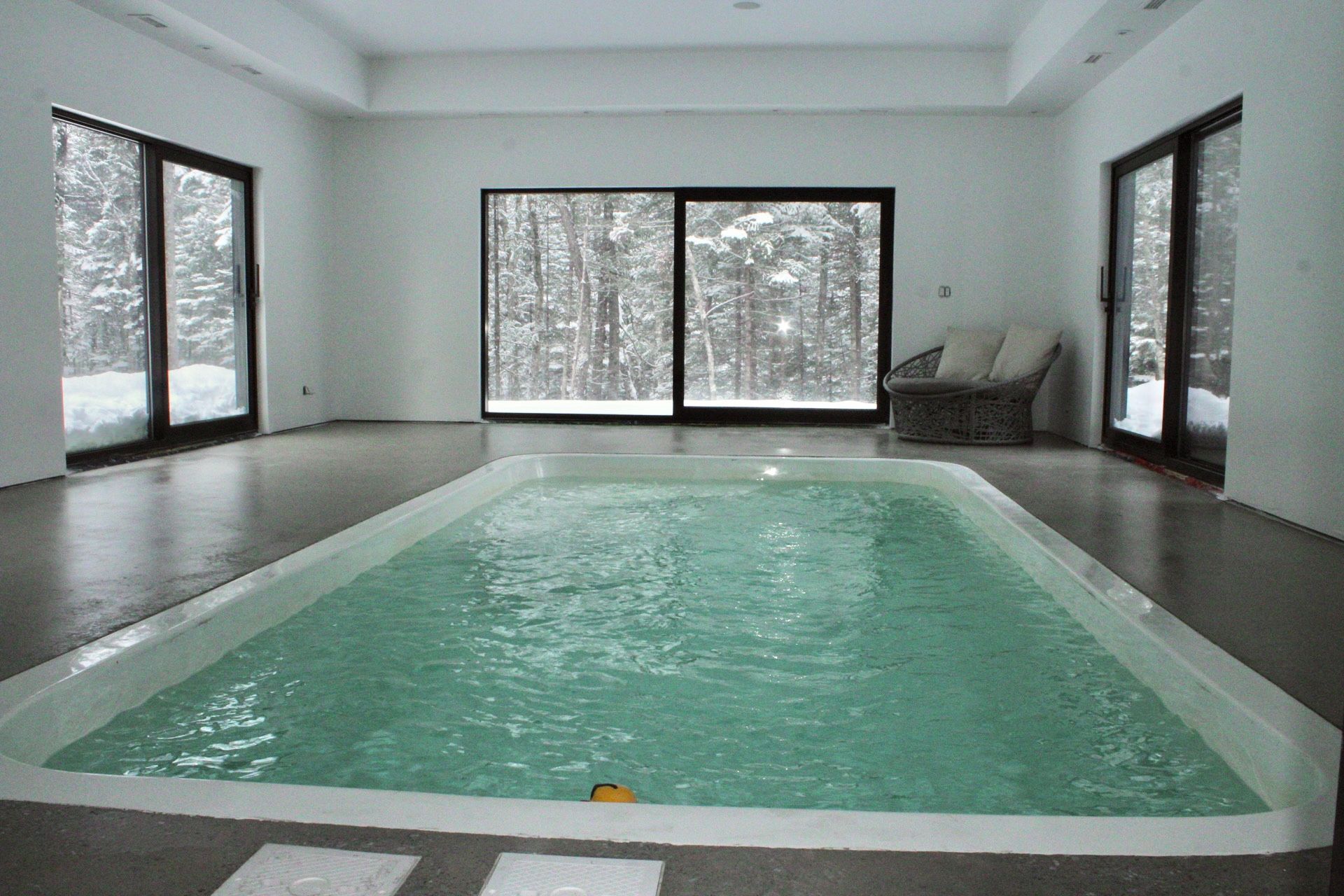 Piscine intérieure avec de grandes baies vitrées donnant sur une forêt enneigée. Murs blancs, sol gris, fauteuil en osier.