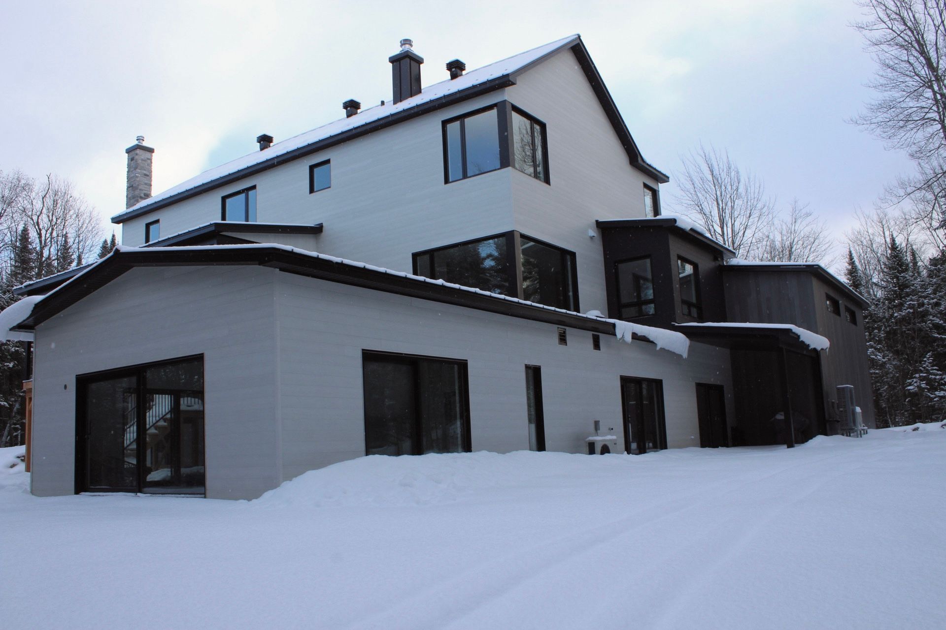 Maison moderne à deux étages en hiver ; sol enneigé. Revêtement gris, toit et encadrements de fenêtres foncés.