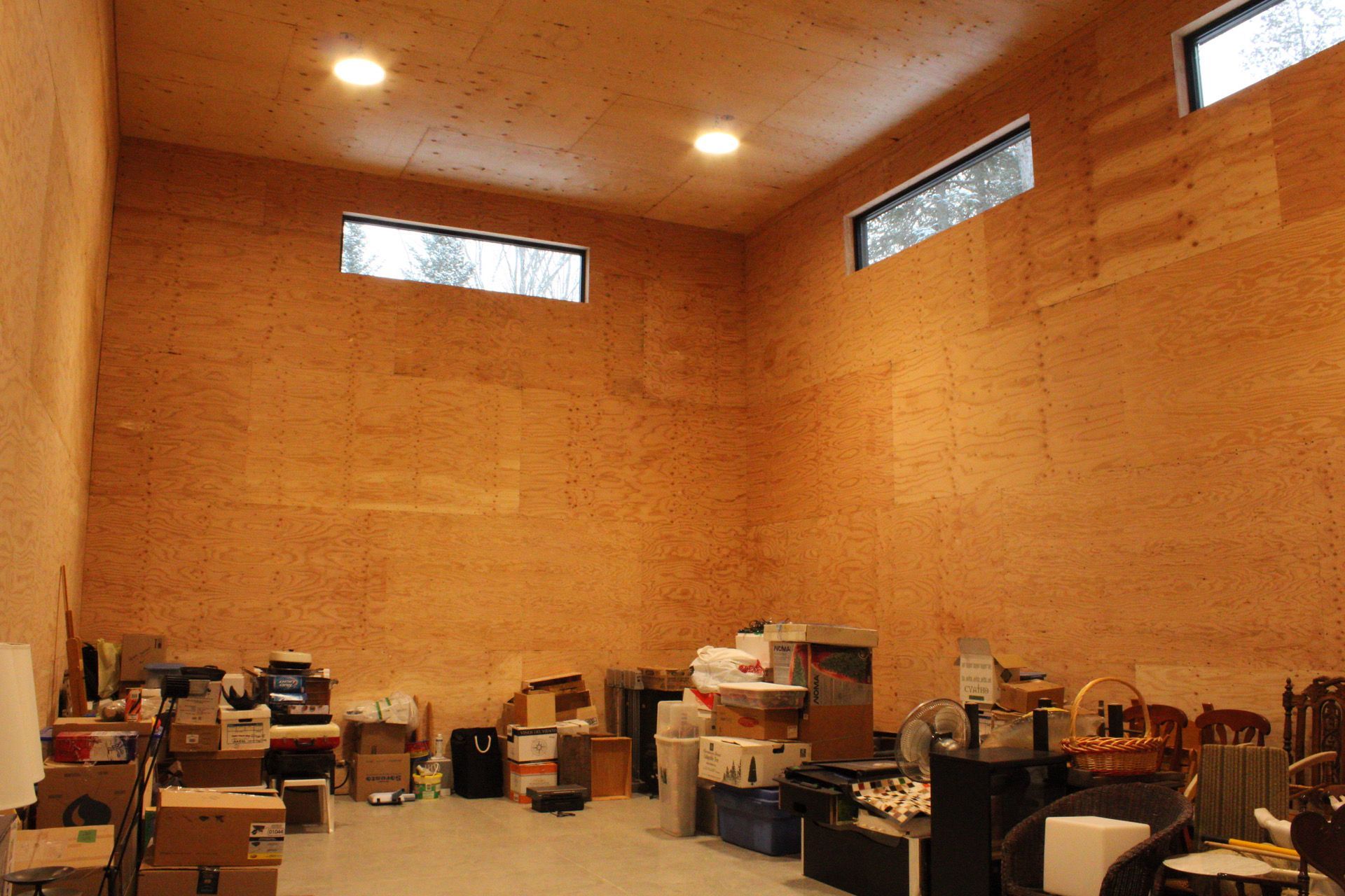 Intérieur d'une pièce avec des cartons et des objets divers contre des murs en panneaux de bois. Deux petites fenêtres, deux plafonniers.