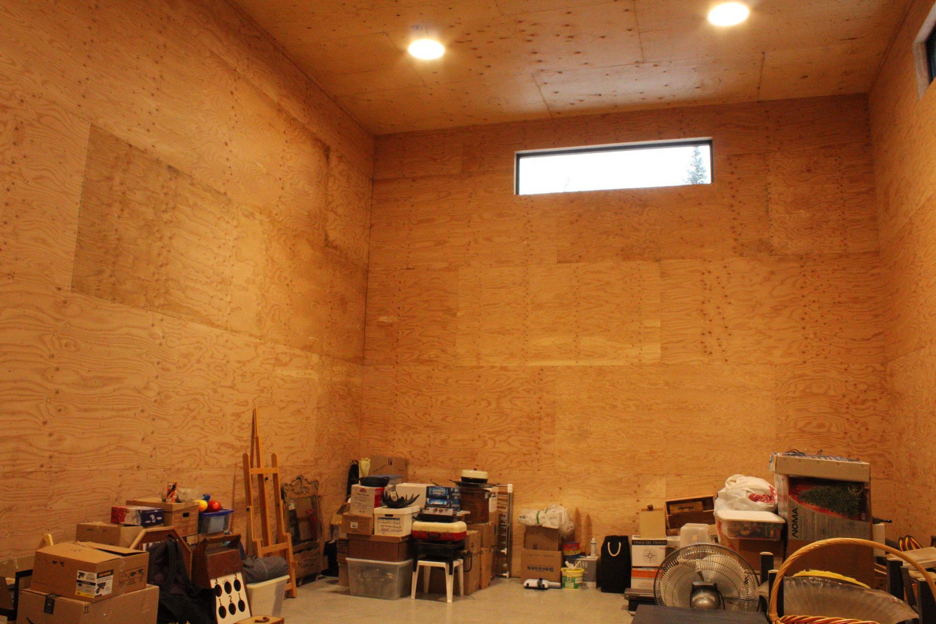 Un débarras encombré avec des murs beiges, une petite fenêtre et des plafonniers. Des cartons et des objets jonchent le sol.