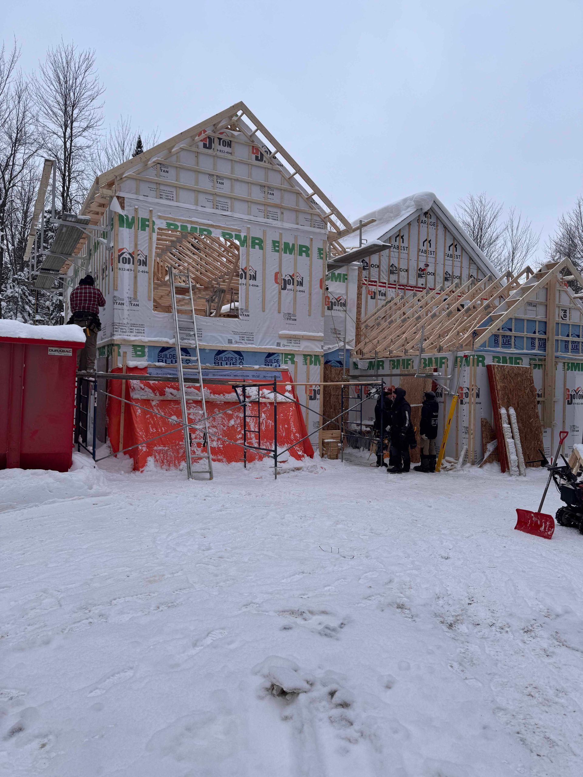 Maison en construction dans un décor enneigé. Ouvriers sur échafaudage, bâche orange, conteneur rouge.