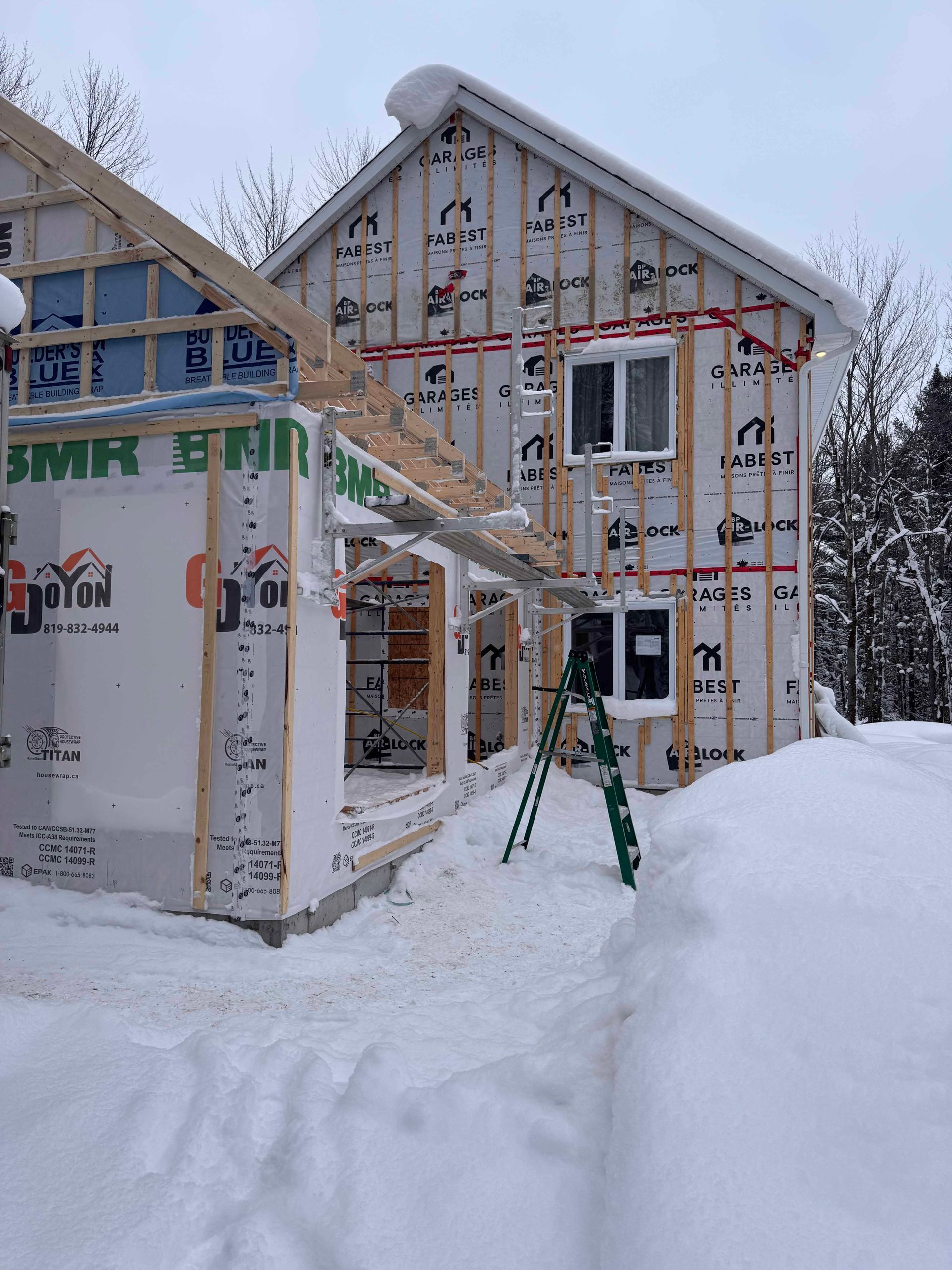 Maison en construction dans un décor enneigé; ossature bois, bardage blanc, échelle et neige au sol.