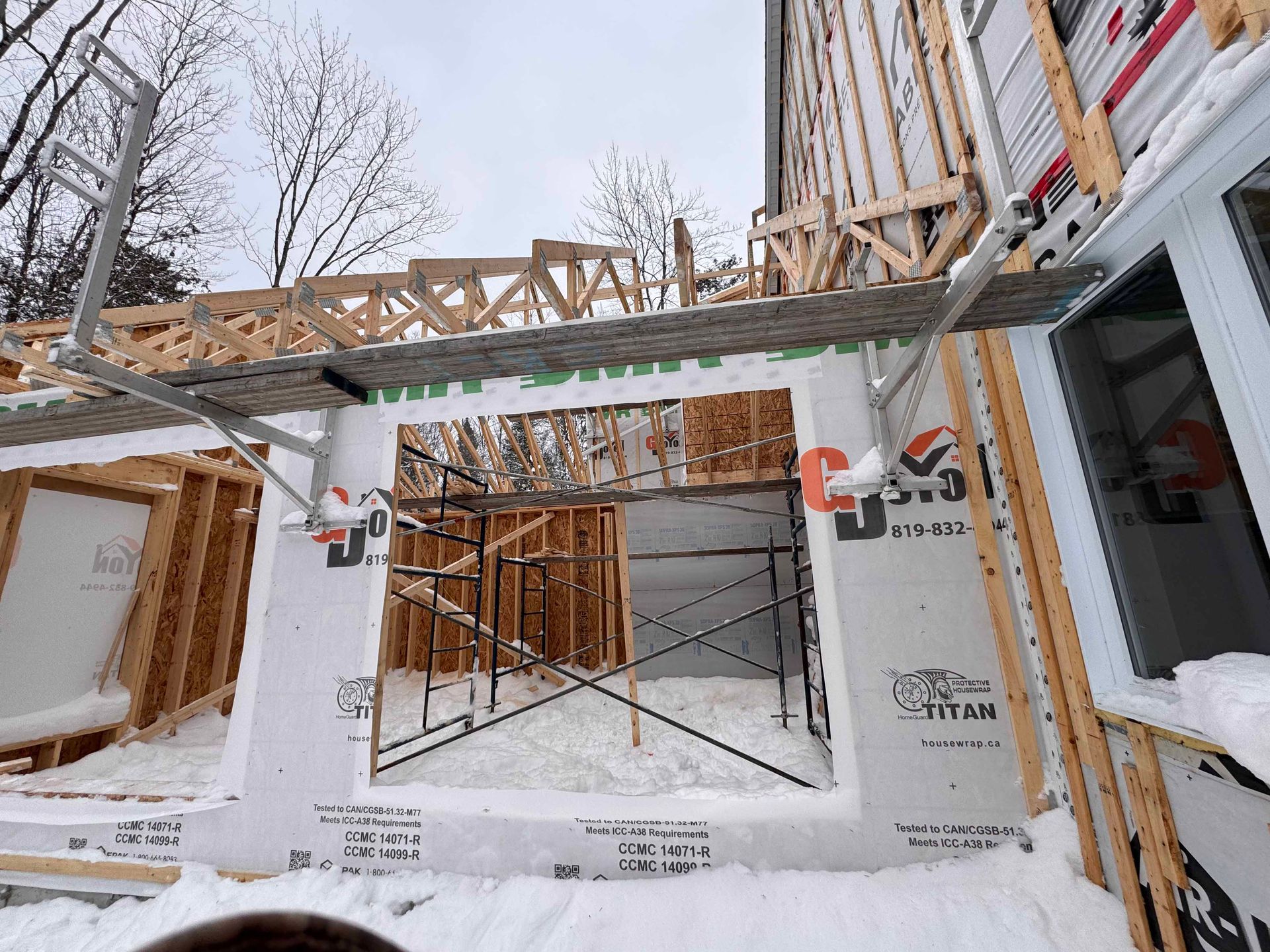 Chantier de construction avec charpente apparente, échafaudages et neige; journée nuageuse.