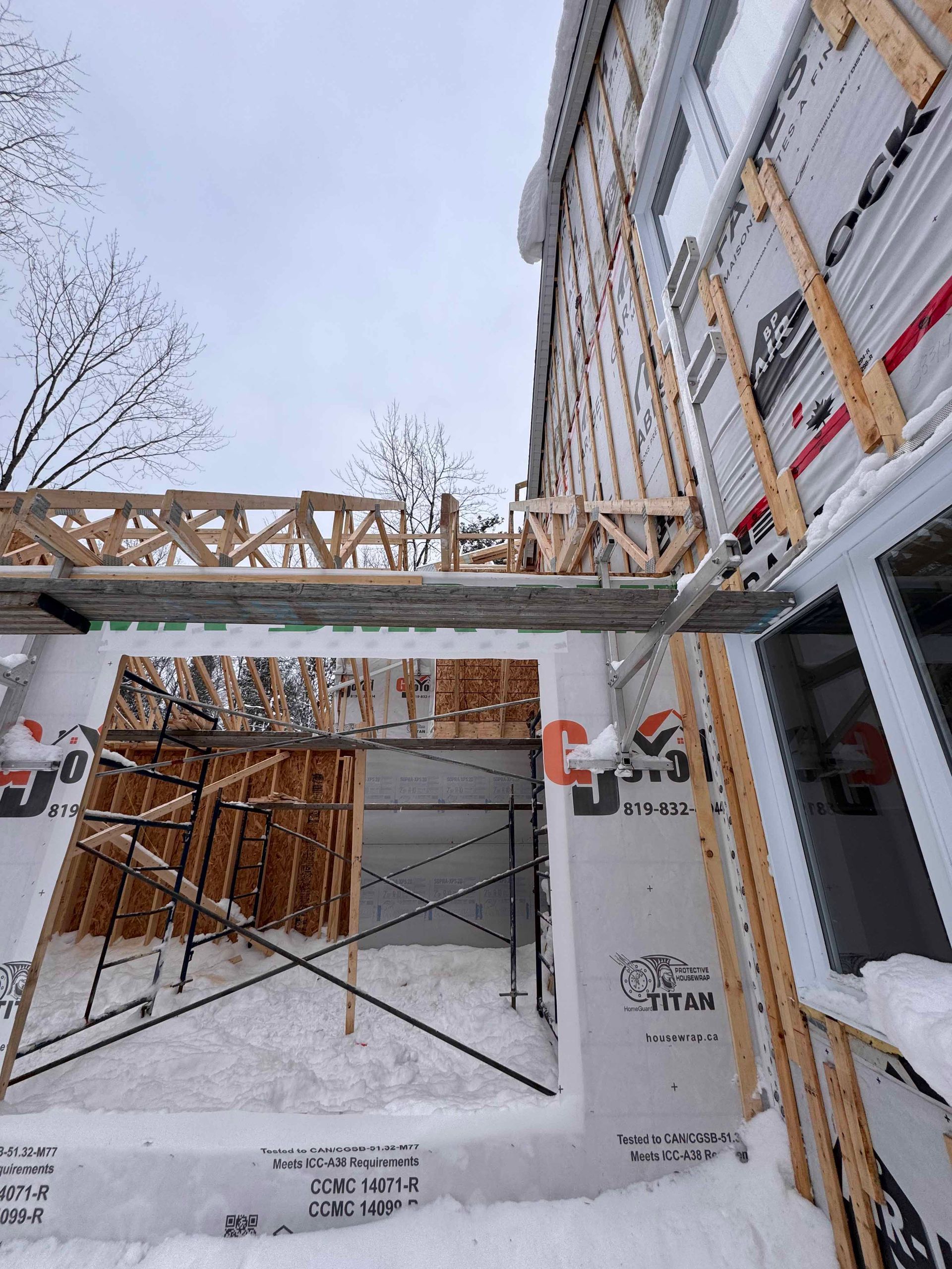 Chantier recouvert de neige, bâtiment partiellement construit avec charpente et échafaudages apparents.
